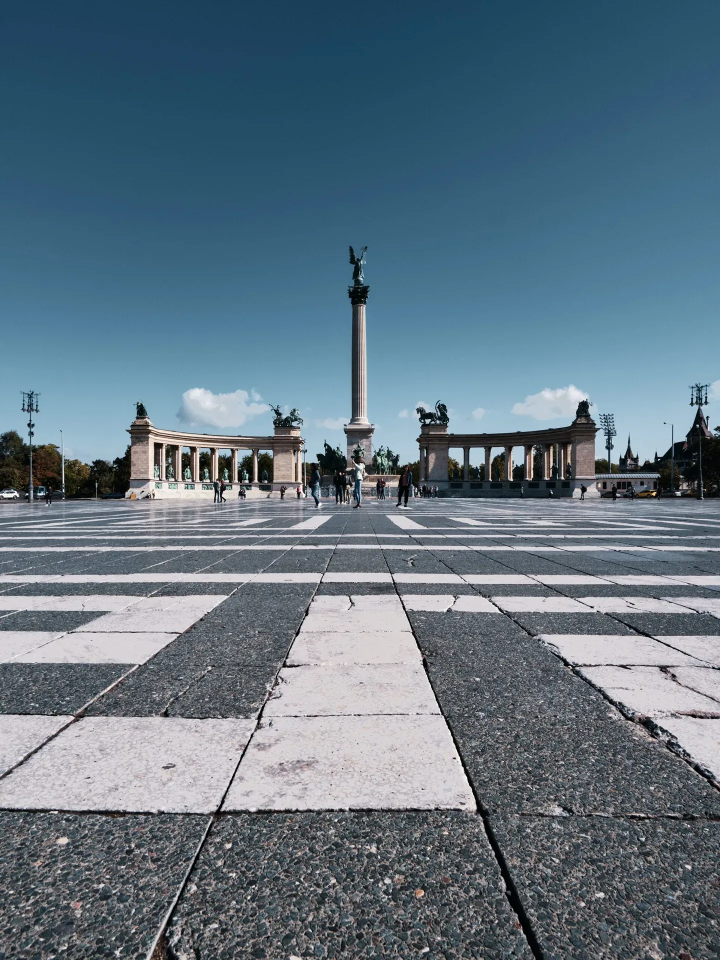 Wide low-angle view of Heroes' Square in Budapest with its central column, semicircular colonnades and checkered pavement under a clear sky.