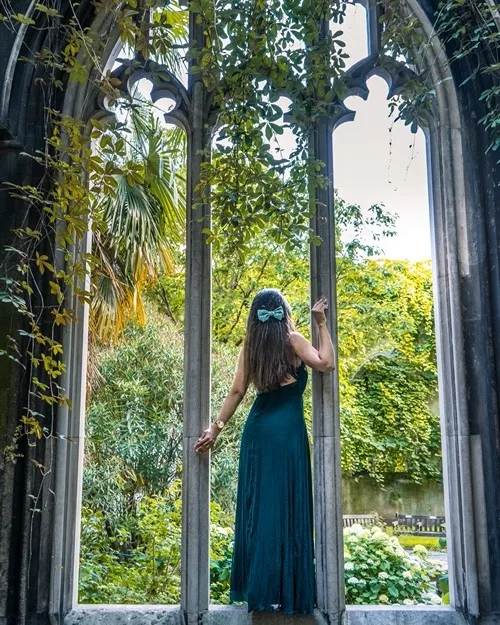 Woman in a teal dress with a matching hair bow standing in a gothic stone window frame, looking out over a lush, overgrown garden.