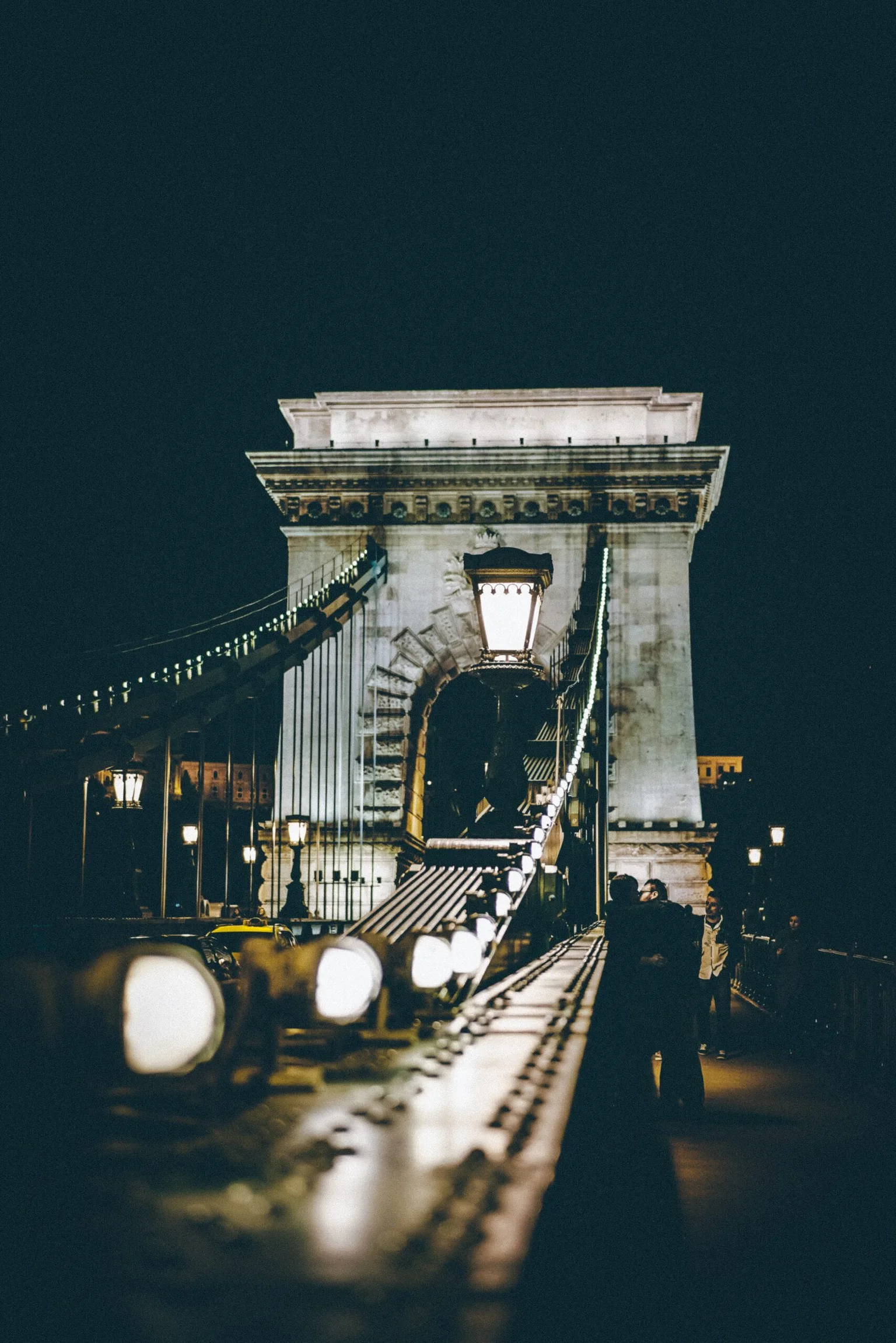 Night view of a lit suspension bridge with stone arches and pedestrians walking along its illuminated balustrade.