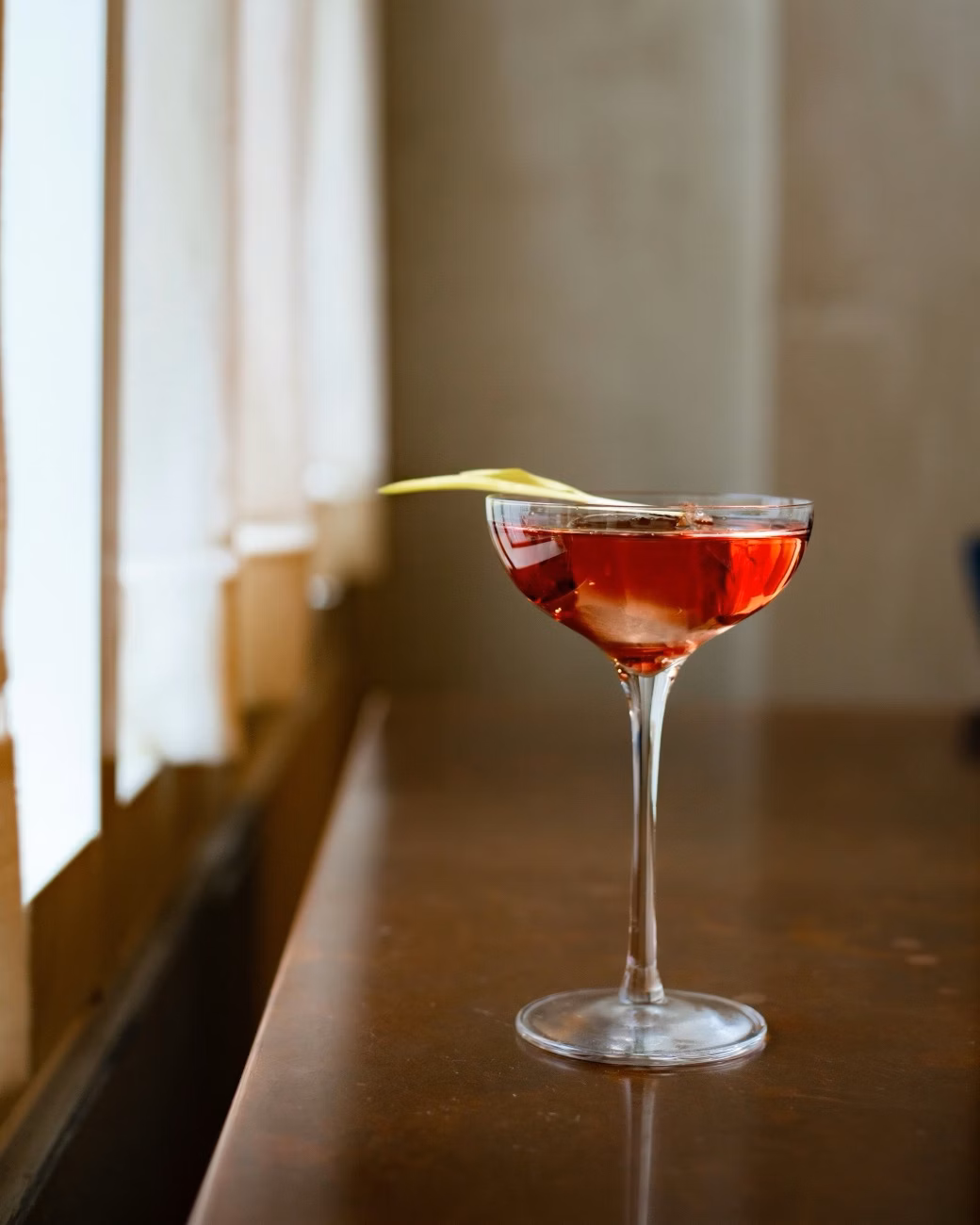 A red cocktail in a stemmed glass with a citrus garnish on a bar by a window