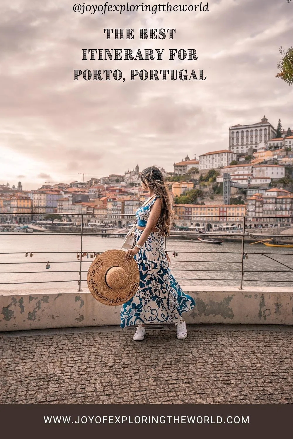 Woman in a blue patterned dress holding a wide straw hat as she overlooks the Ribeira waterfront and colourful hillside houses in Porto, Portugal.