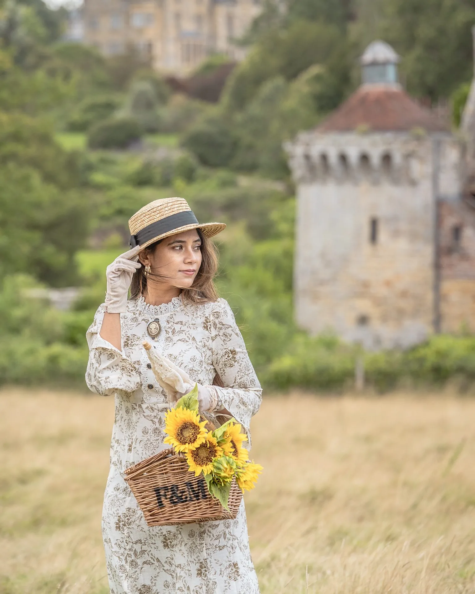 Young woman in a vintage floral dress and straw boater hat holds a wicker basket of sunflowers in a meadow with a stone turreted manor in the background.