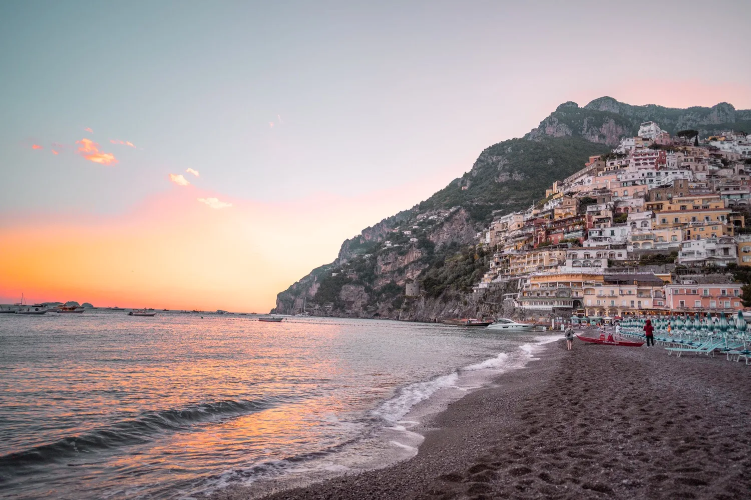 Colourful cliffside town with stacked pastel buildings beside a pebble beach and calm sea at sunset.