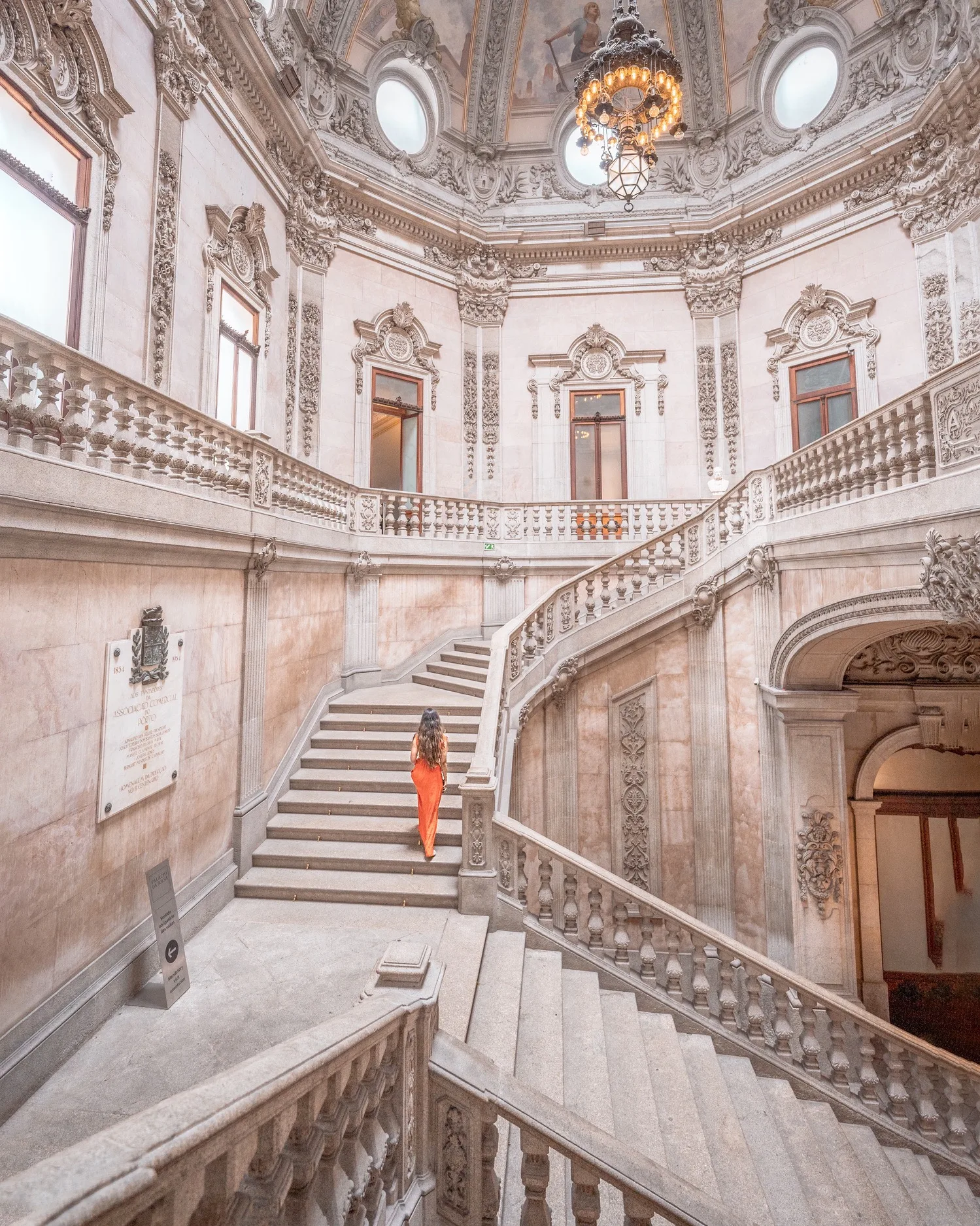 Woman in a red dress ascending an ornate marble staircase inside a grand neoclassical interior with carved balustrades and a domed ceiling chandelier.