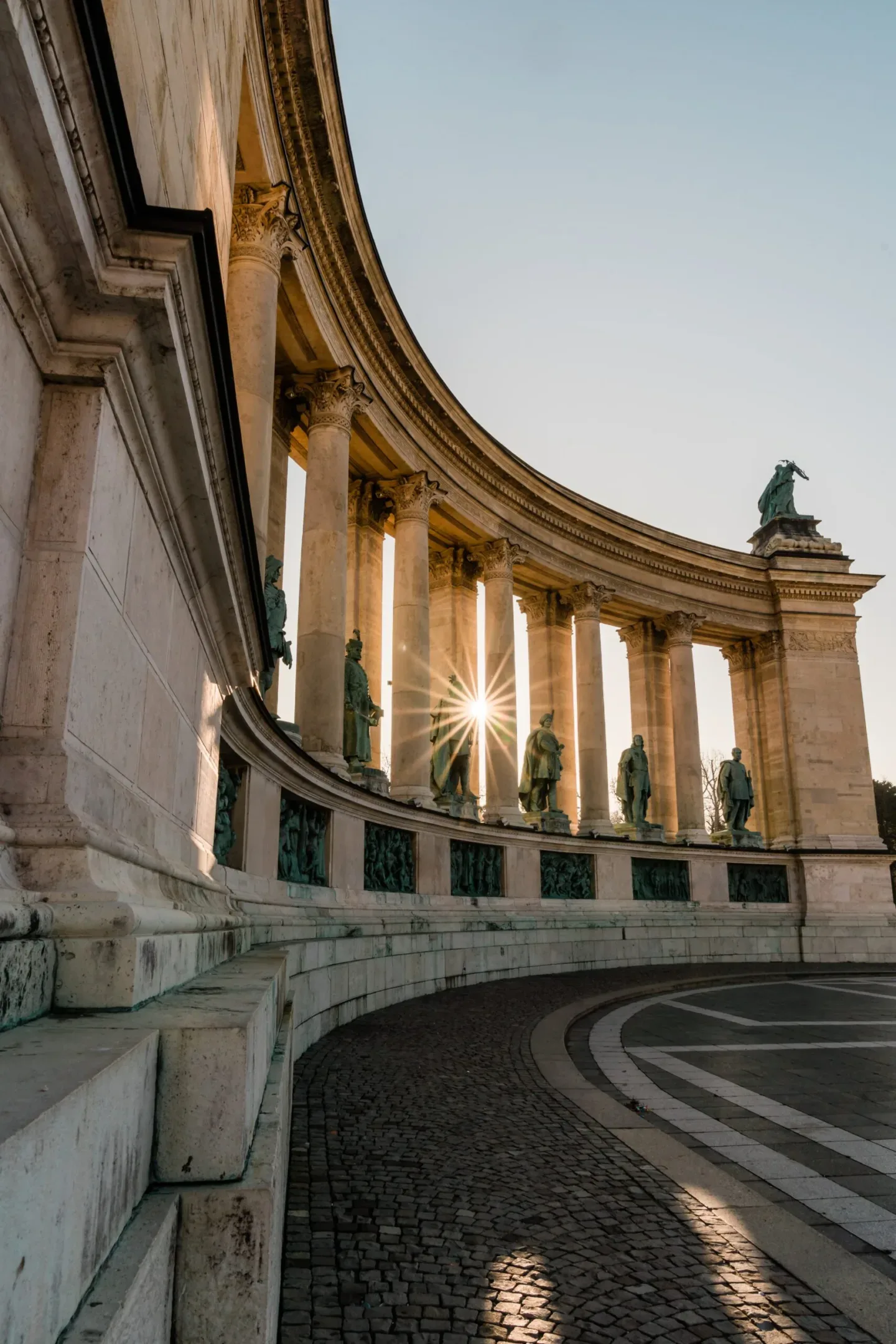 Curved colonnade and bronze statues of Heroes' Square in Budapest lit by a low sunburst between the columns.