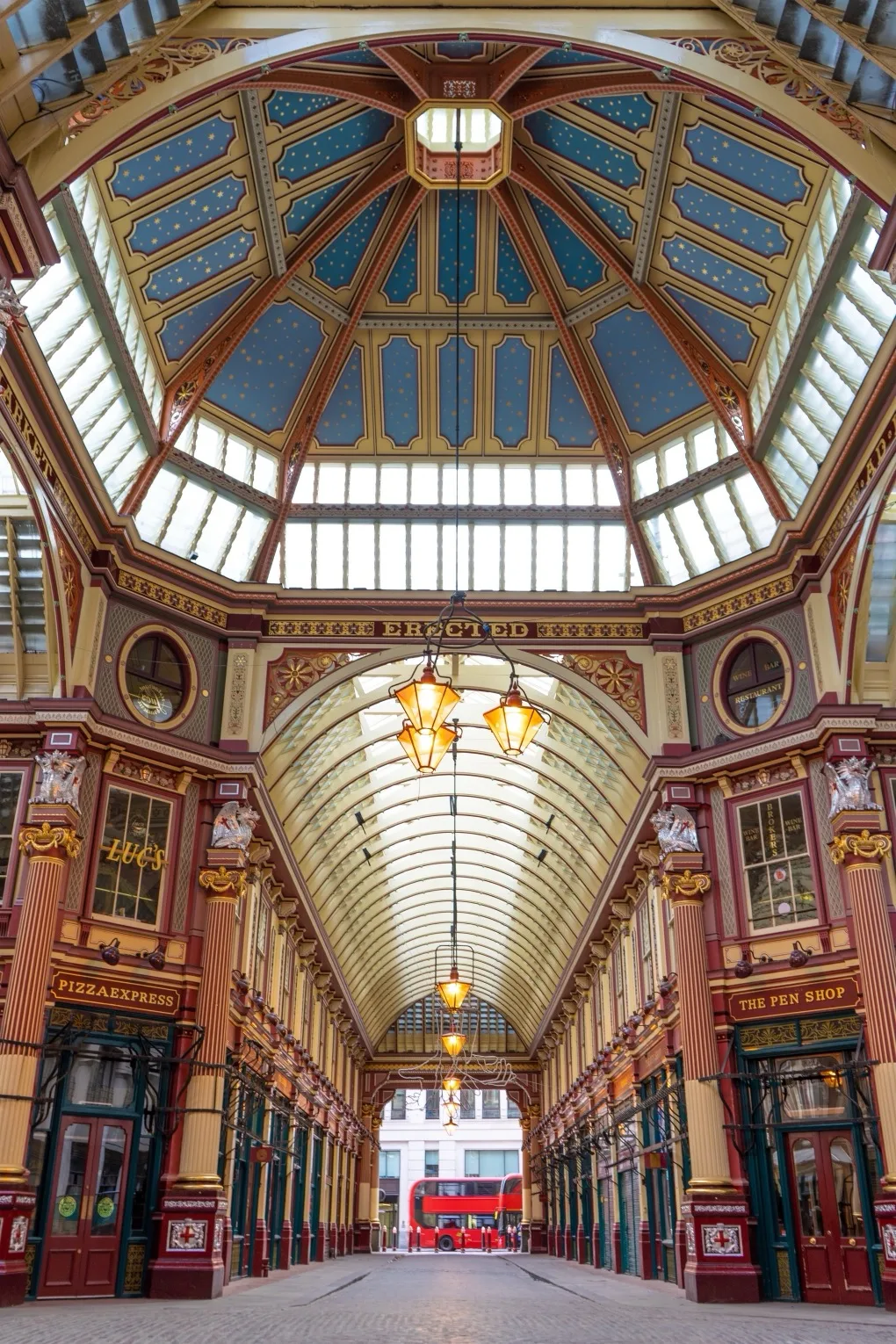 Victorian covered arcade with a vaulted glass roof, ornate painted dome and rows of shops leading to a red double-decker bus.