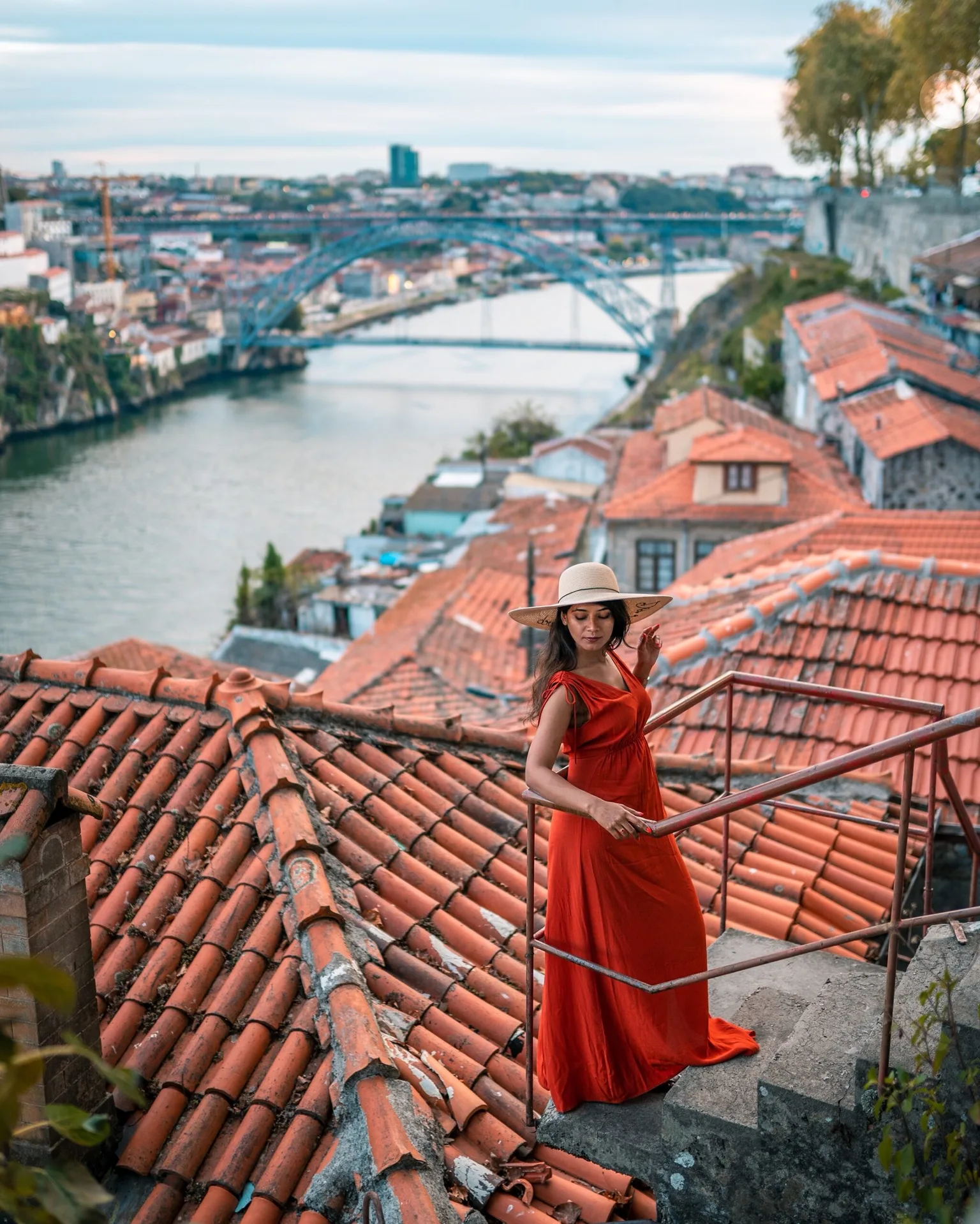 Woman in a red dress and wide-brimmed hat standing on stone steps above terracotta rooftops with a river and arched bridge in the background.