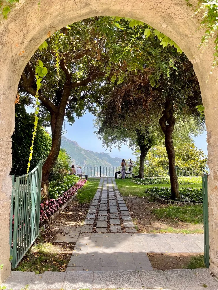 Stone archway framing a tree-lined garden path leading to a terrace with people overlooking distant mountains and a blue sky.