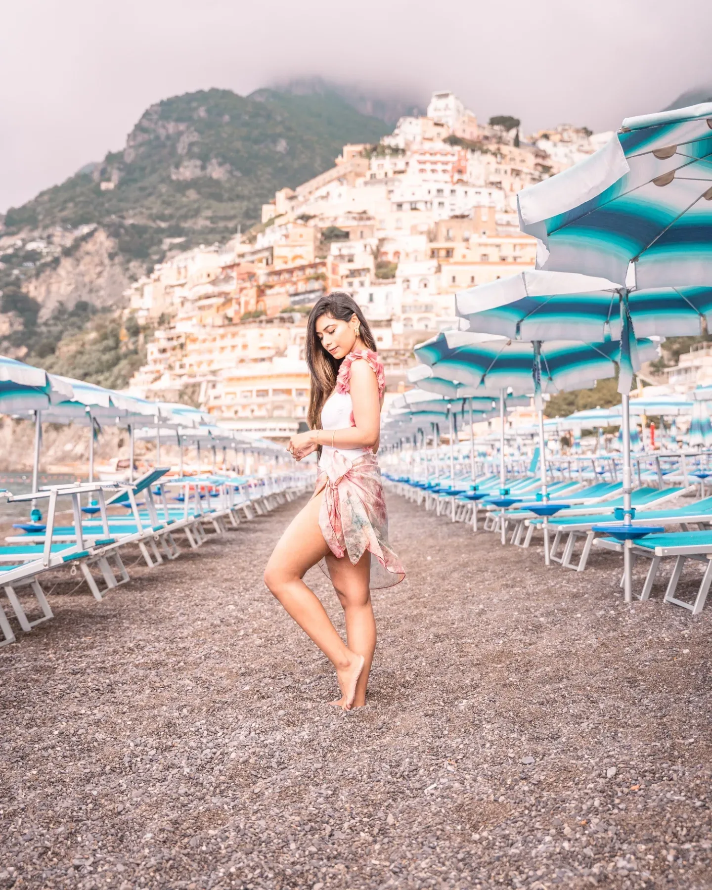 Woman in a light sarong standing barefoot between rows of blue beach umbrellas with cliffside village in the background.