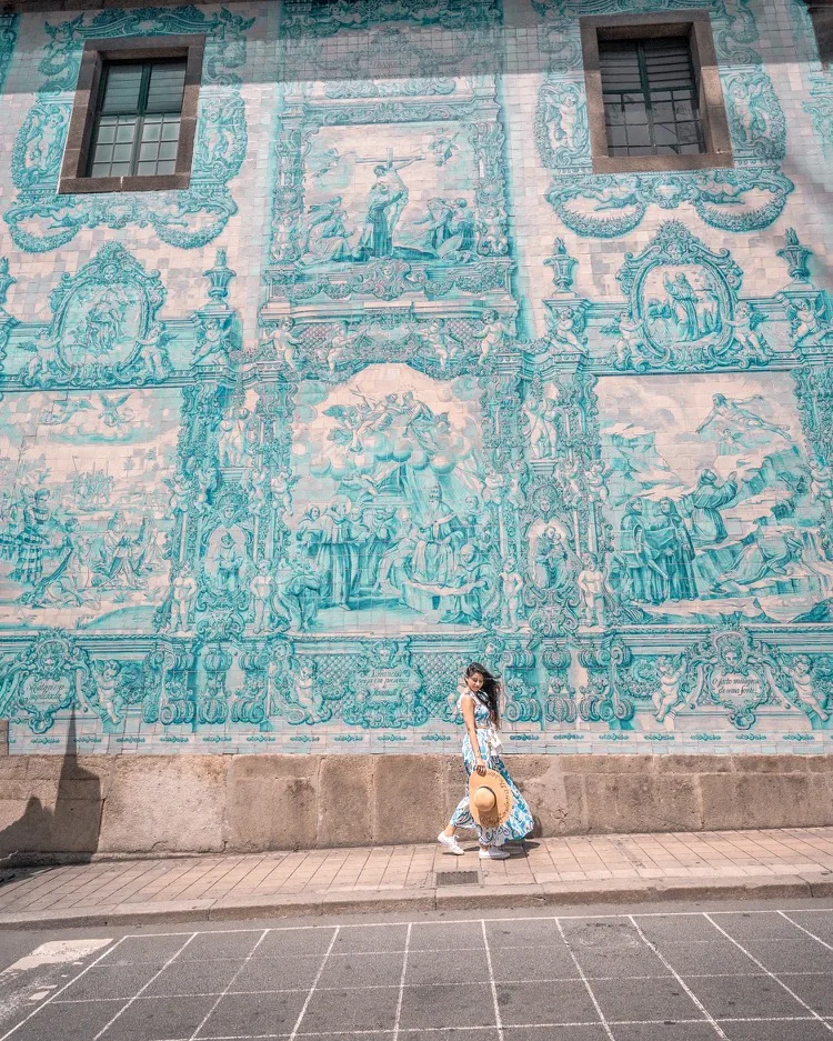 Woman in a flowing dress and sunhat walking past a large blue azulejo-tiled mural on a city street.