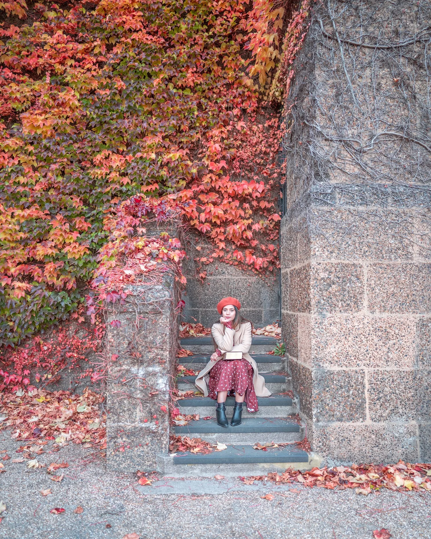 Woman in a red beret and autumn dress sitting on stone steps framed by vivid red and orange ivy on a city wall.