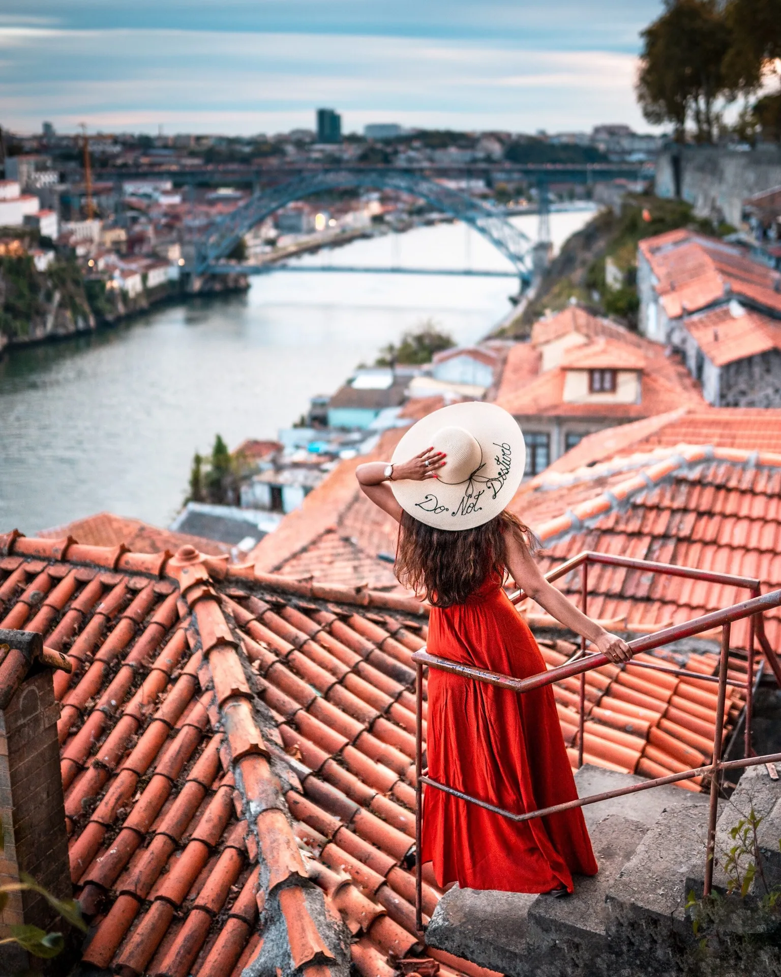 Woman in a red dress and wide-brimmed hat overlooking terracotta rooftops and a river with an arched bridge in the distance.