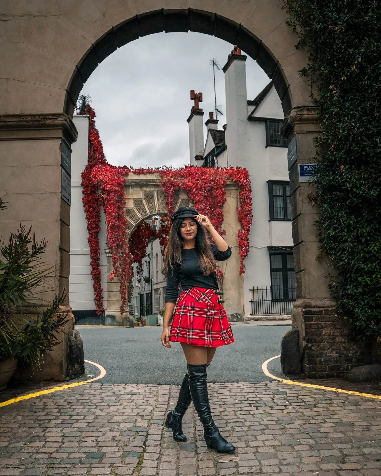 Woman in a red tartan skirt and black over-the-knee boots posing beneath an arched gateway draped with red ivy in a cobbled street.