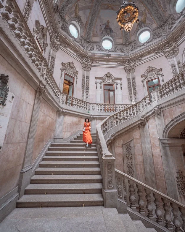 Woman in a flowing orange dress descending an ornate marble staircase beneath a domed ceiling with decorative plasterwork and a central chandelier
