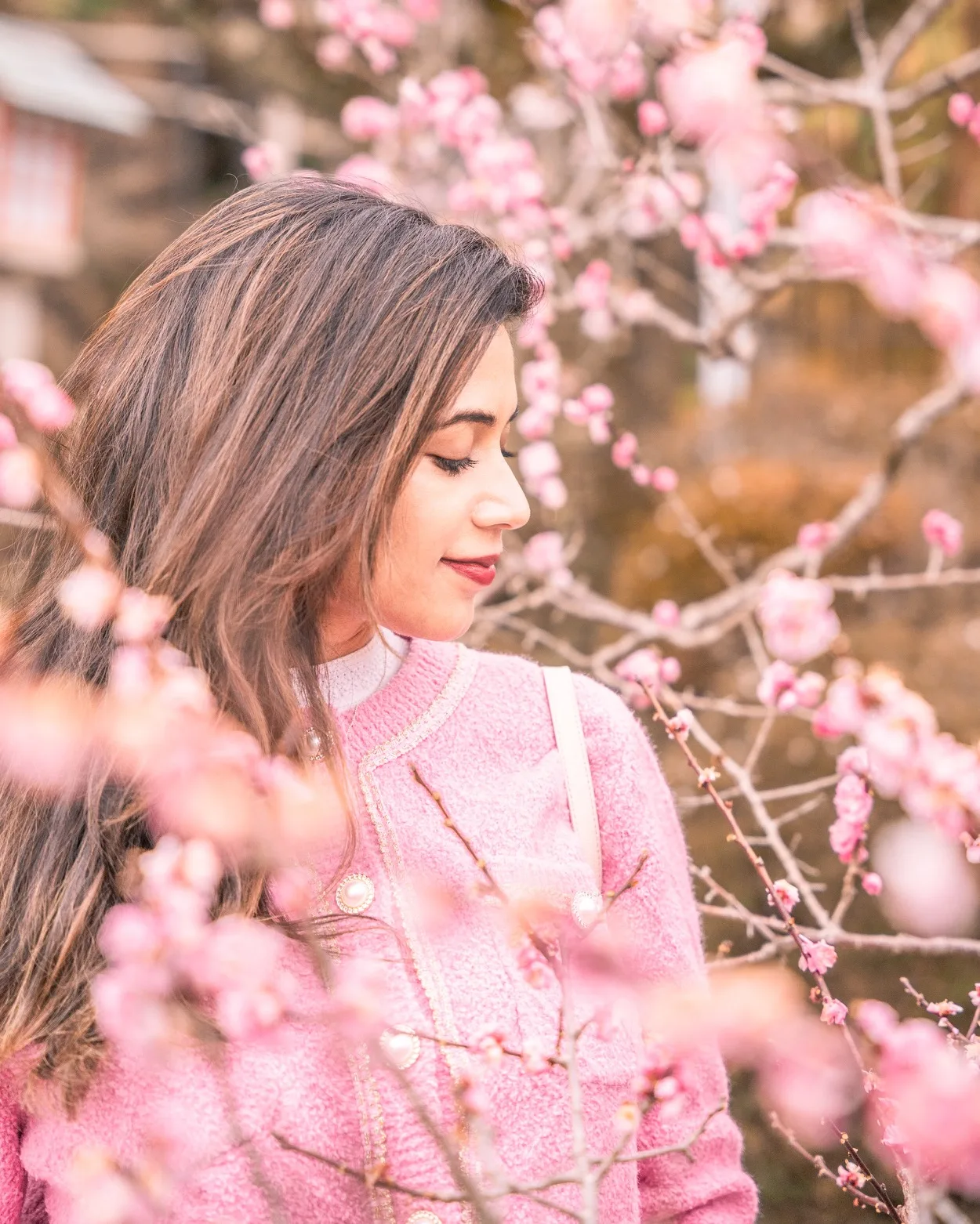 Young woman in a pink coat standing among blooming pink cherry blossoms, smiling softly with eyes closed in Tokyo, Japan.