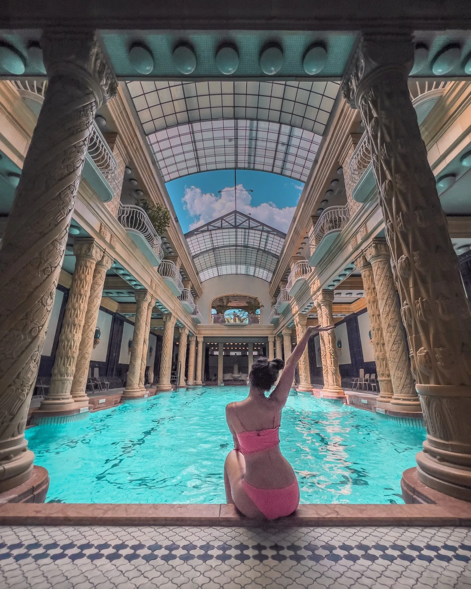 Woman in a pink bikini sitting at the edge of an ornate indoor pool framed by carved columns and a glass atrium ceiling.