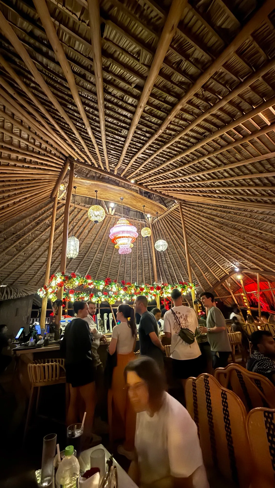 Crowded tropical bar under a high thatched roof with hanging lanterns and a flower-decorated central serving area.