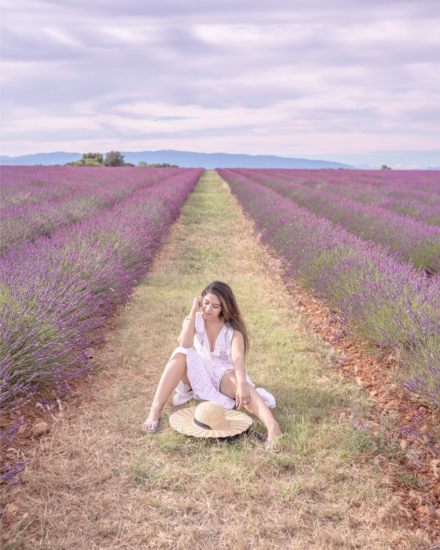 Woman in a white summer dress sitting on a grassy path among rows of purple lavender fields, straw hat beside her.