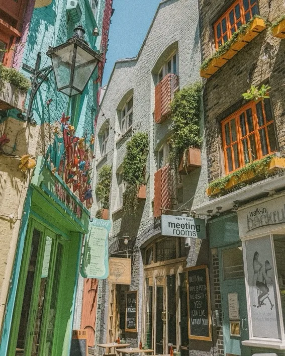 Narrow sunlit alley lined with colourful shopfronts, hanging window boxes and a sign reading "meeting rooms" above a café entrance.