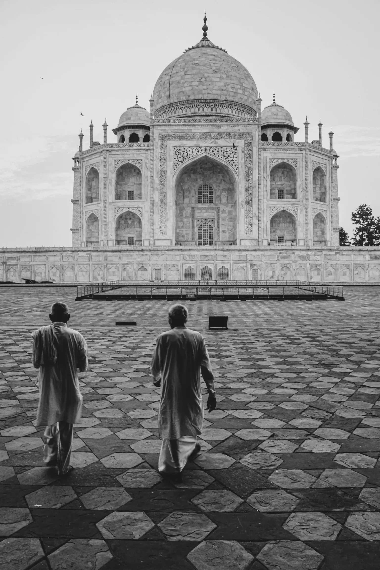 Two men in traditional white clothing walk across patterned stone tiles toward the Taj Mahal.