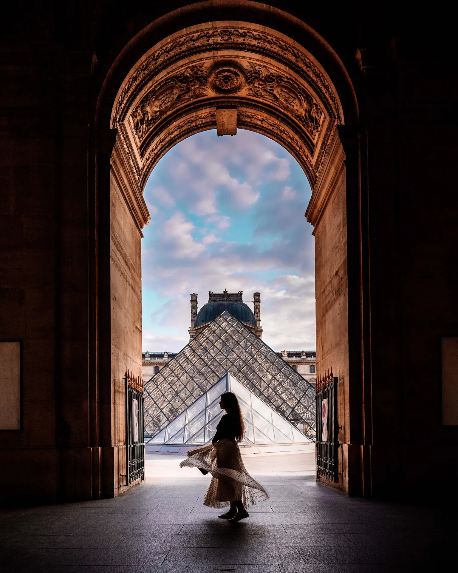 Silhouette of a woman twirling beneath an ornate archway with the Louvre Pyramid and cloudy sky framed beyond.