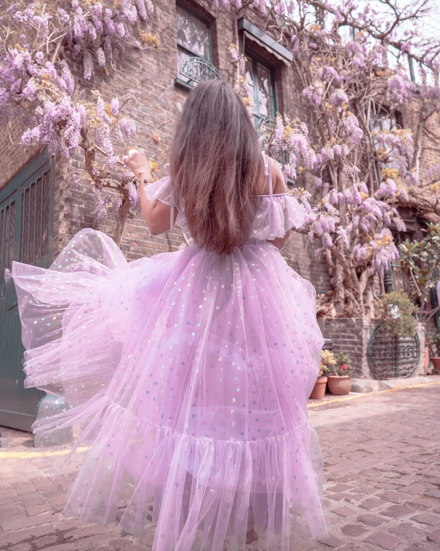 Woman in a flowing lavender tulle dress twirling on a cobbled street lined with wisteria-covered brick buildings.