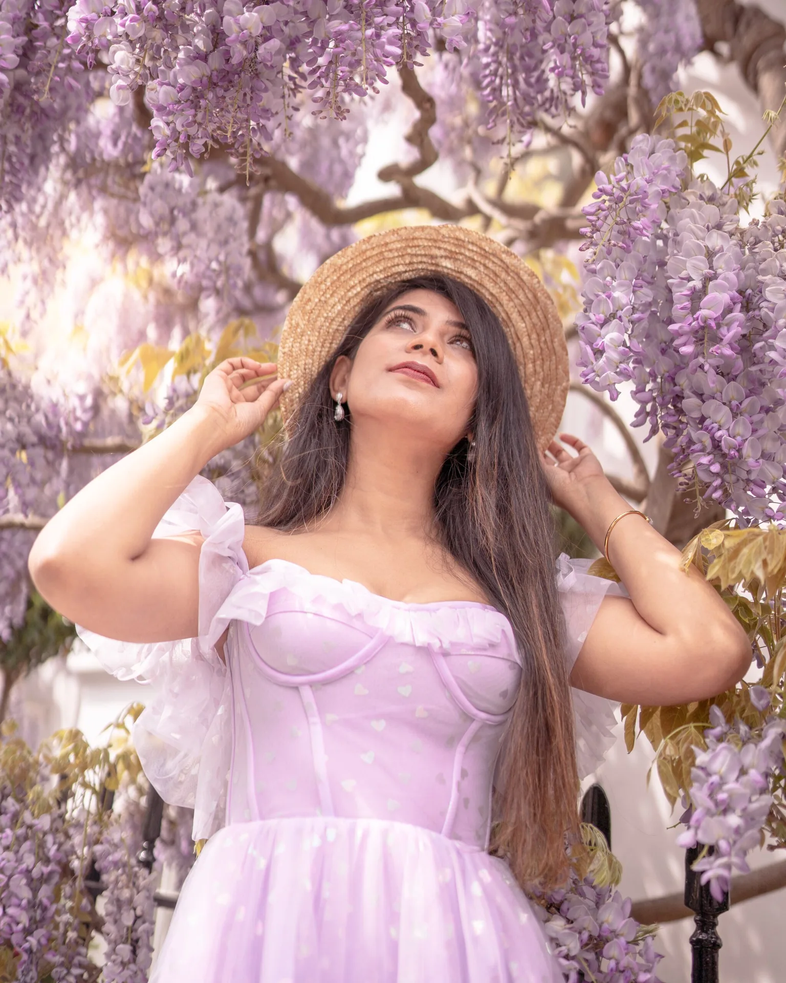 Woman in a lavender dress and straw hat gazes up amid cascading wisteria blossoms.