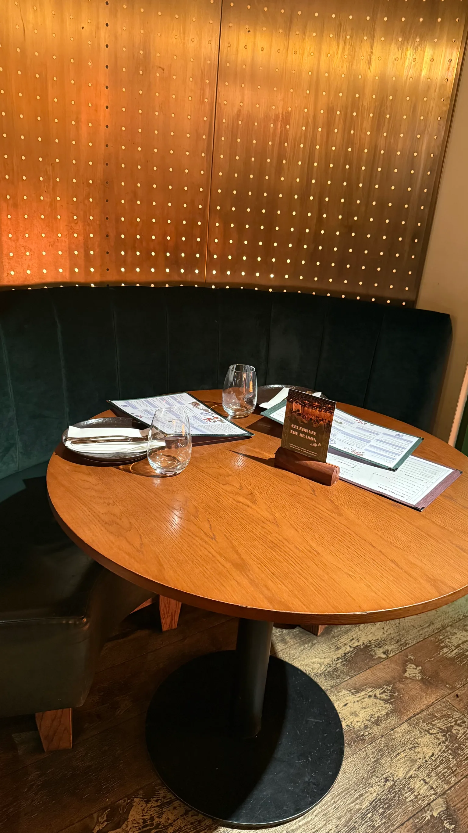 Round wooden restaurant table set with menus, two water glasses, cutlery and a small sign, against a dark upholstered banquette and perforated wooden wall.