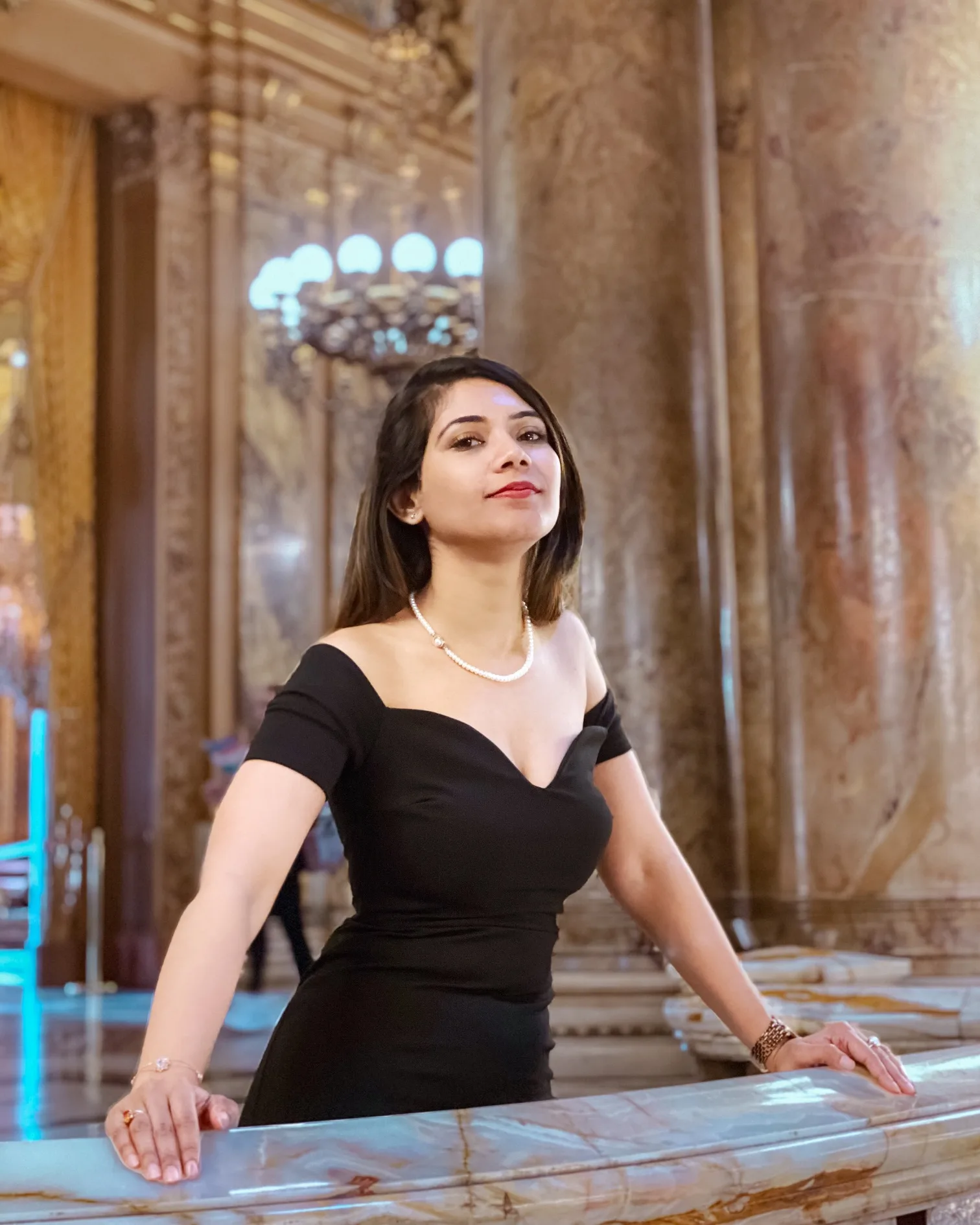 Woman in an elegant black off-shoulder dress and pearl necklace leaning on a marble balustrade inside an ornate, columned interior.