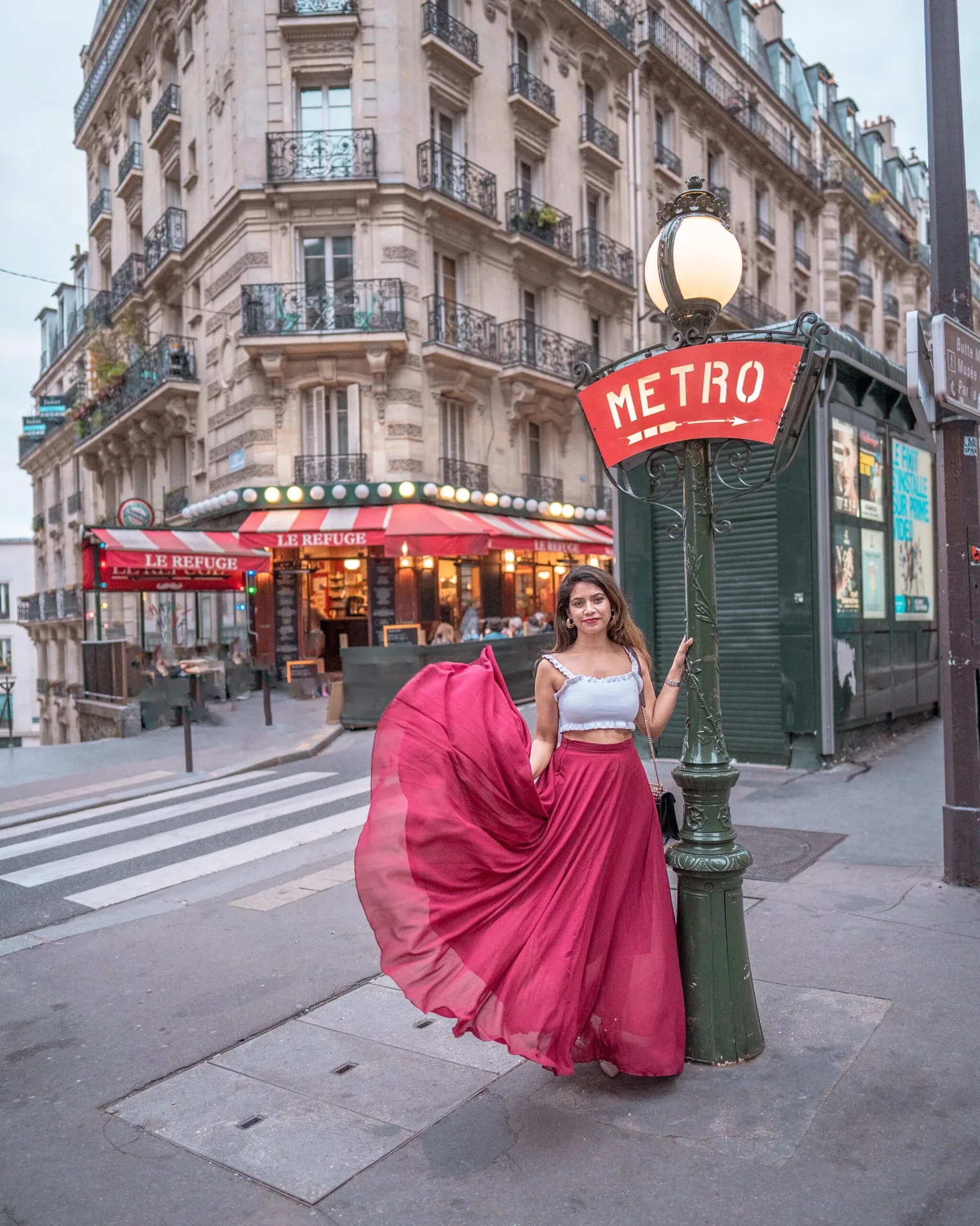Woman in a flowing magenta skirt posing by a vintage "METRO" sign with a Parisian café and Haussmannian buildings in the background.