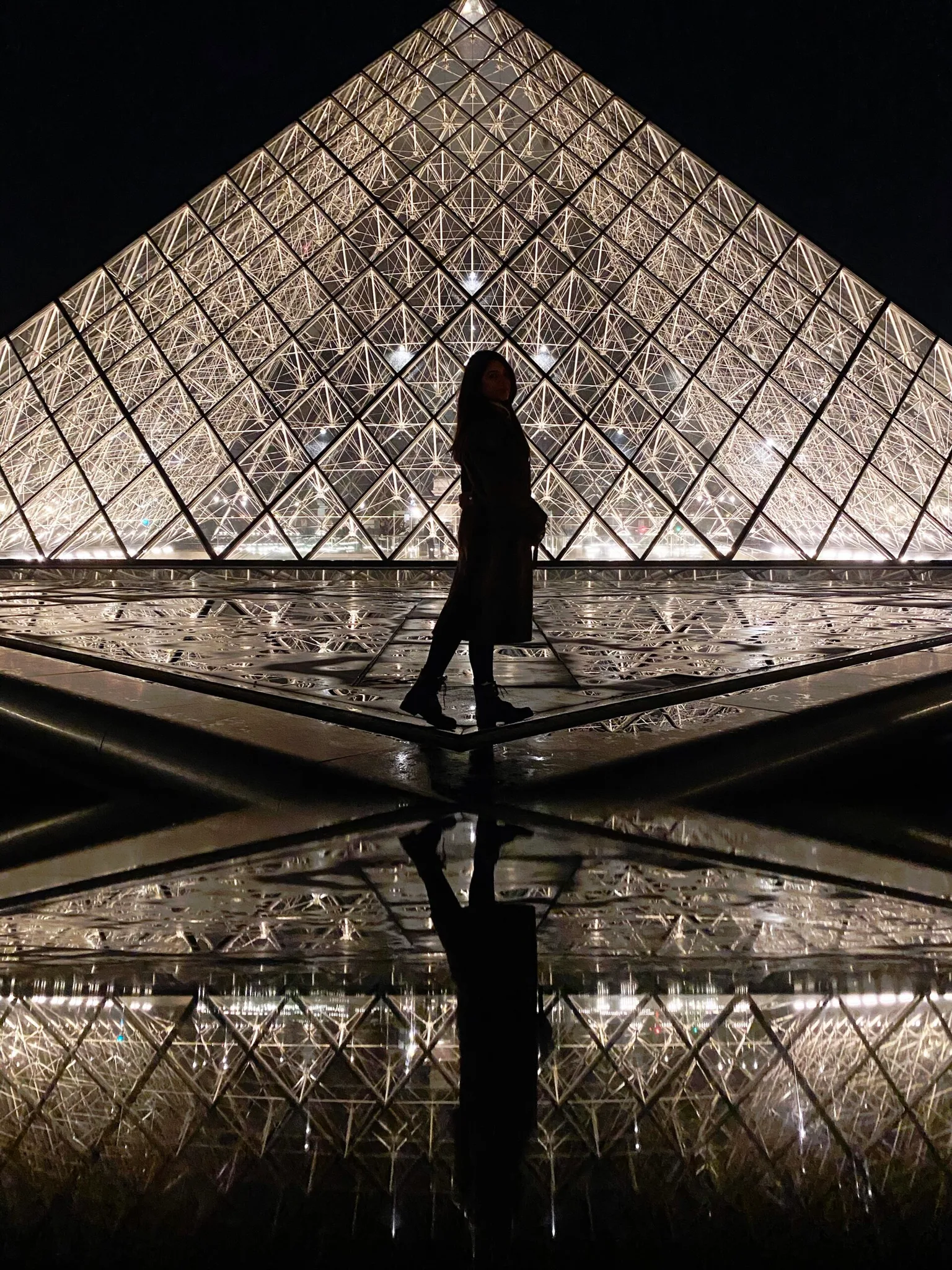 Silhouette of a person standing in front of the illuminated glass pyramid at the Louvre at night, with a perfect reflection in the wet plaza.