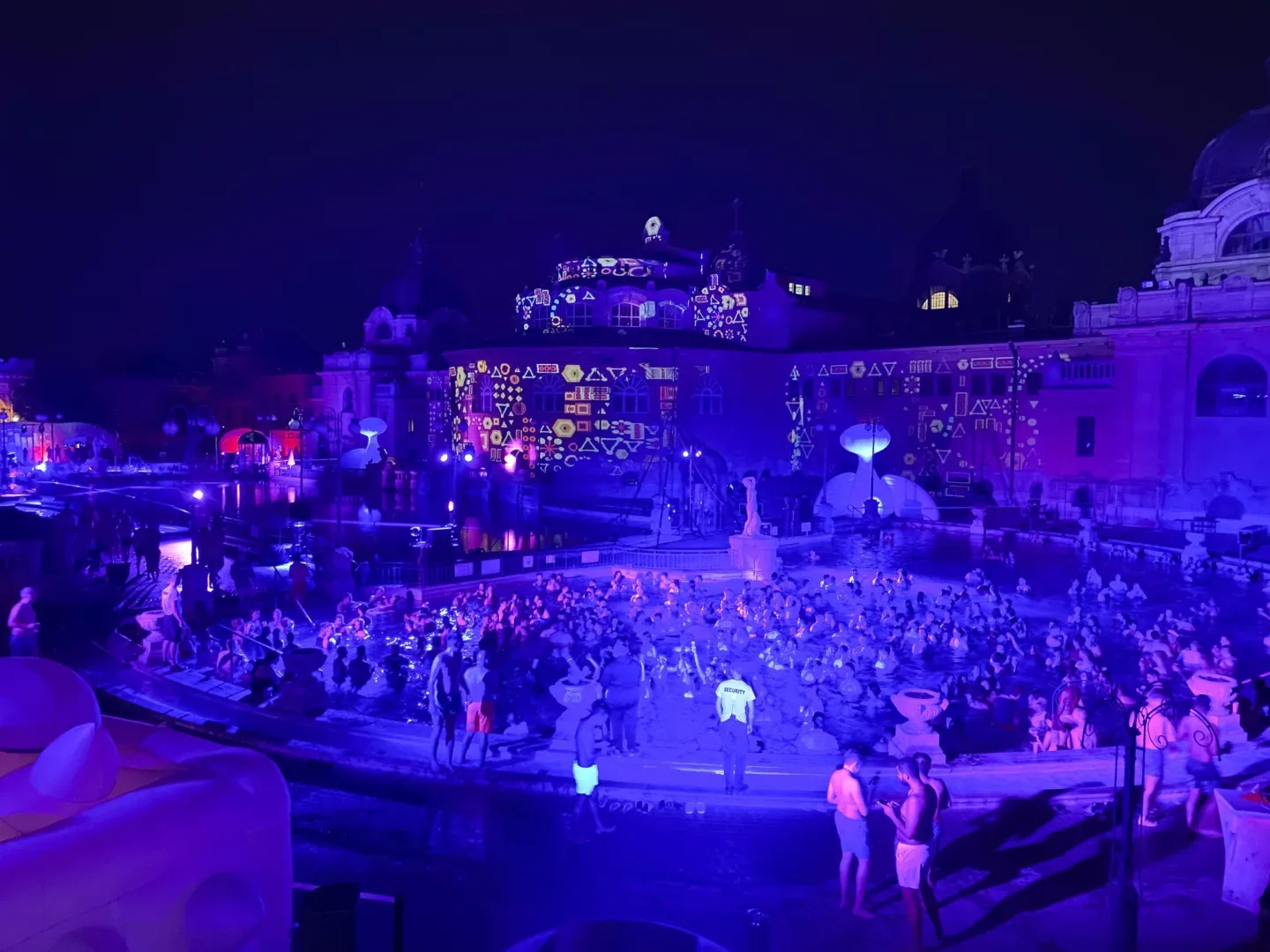 Crowd of people enjoying a night-time light show and pool party at a historic thermal bath complex lit in vivid purple and projected patterns.