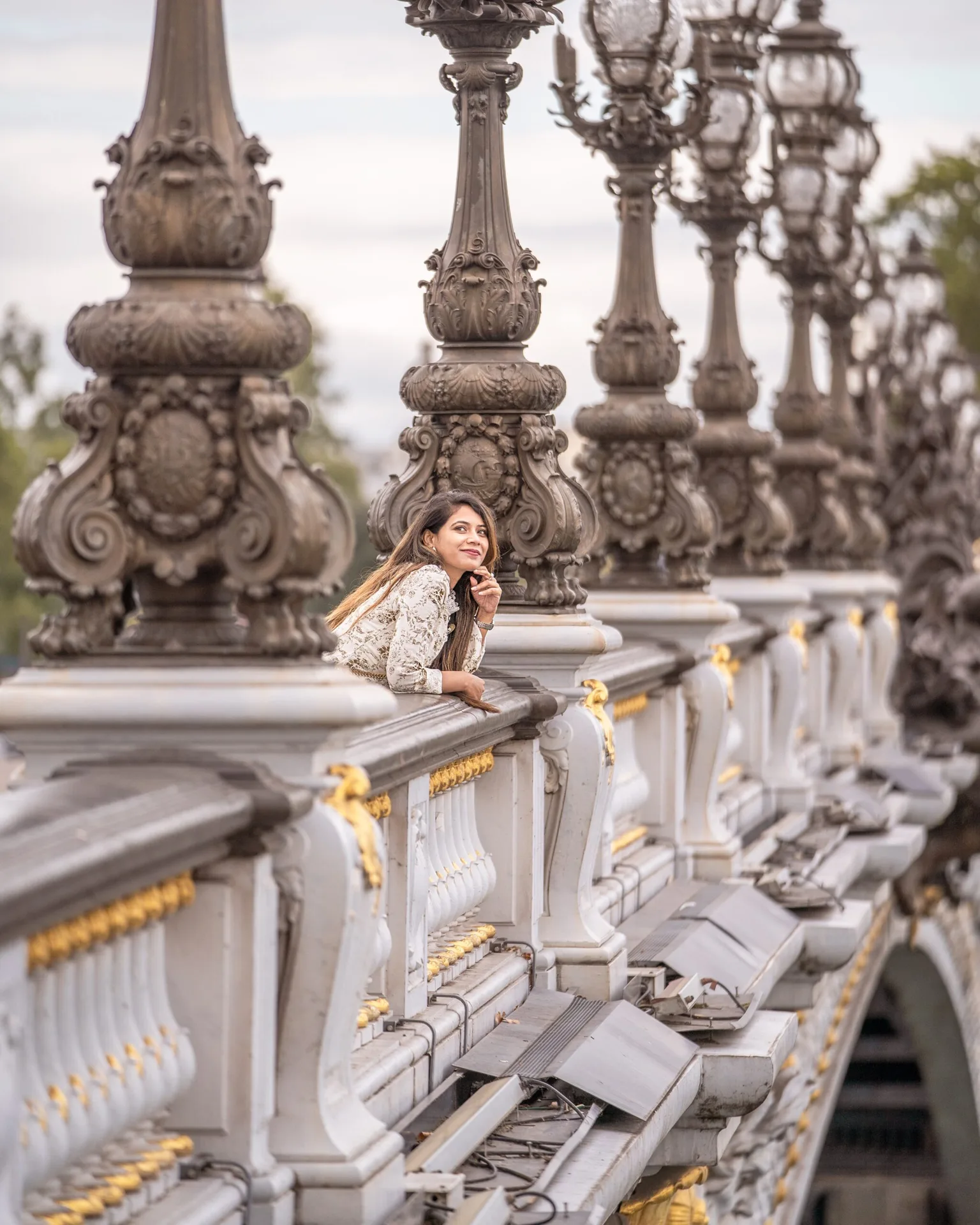 Young woman leaning on ornate white-and-gold bridge balustrade, framed by a row of decorative bronze lamp posts.