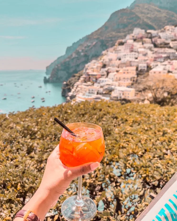 Hand holding a bright orange aperitif in a stemmed glass with straw, overlooking sunlit cliffside houses and boats along a turquoise coastal bay.