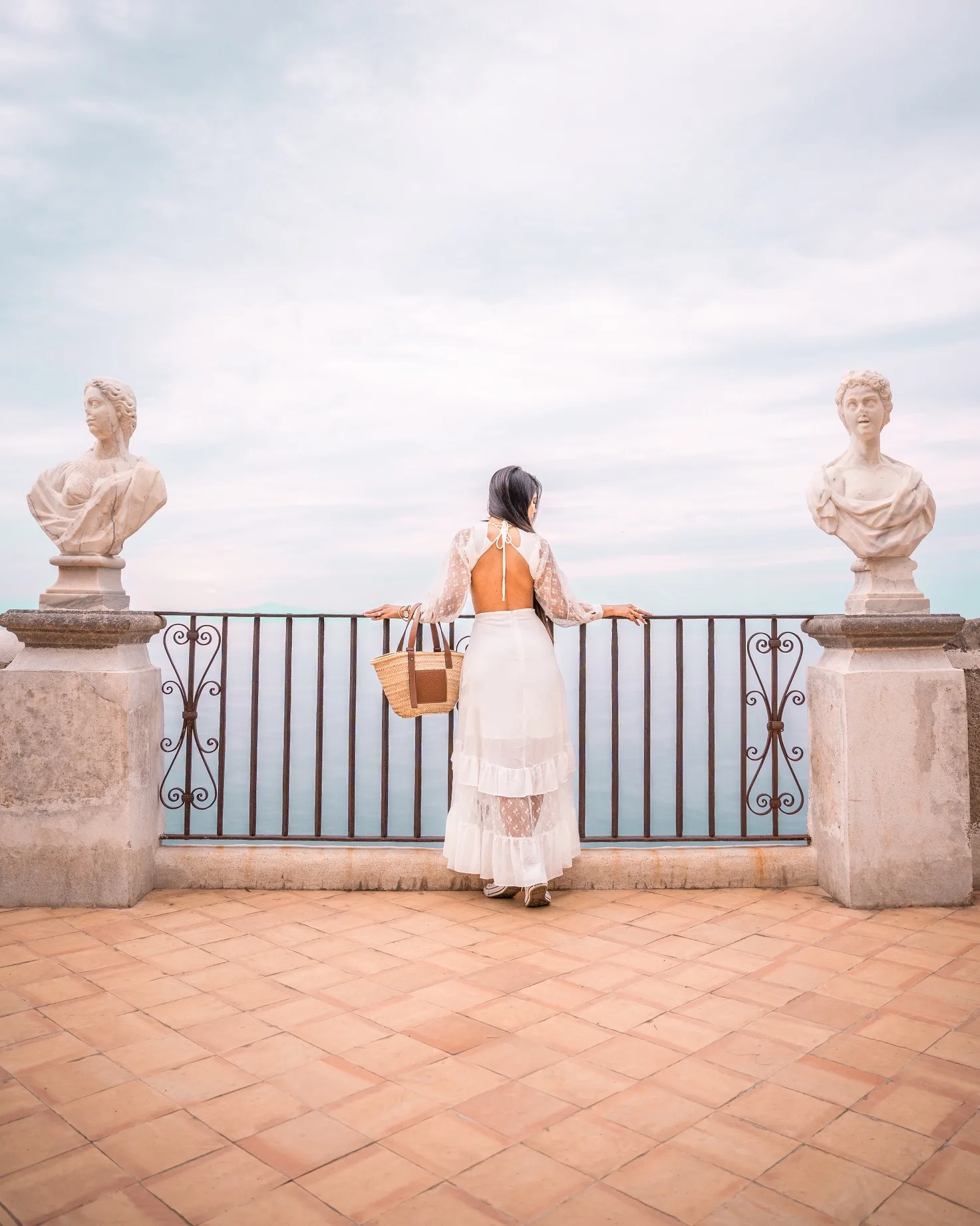 Woman in a white open-back dress holding a straw tote and leaning on a seaside terrace railing flanked by classical busts.