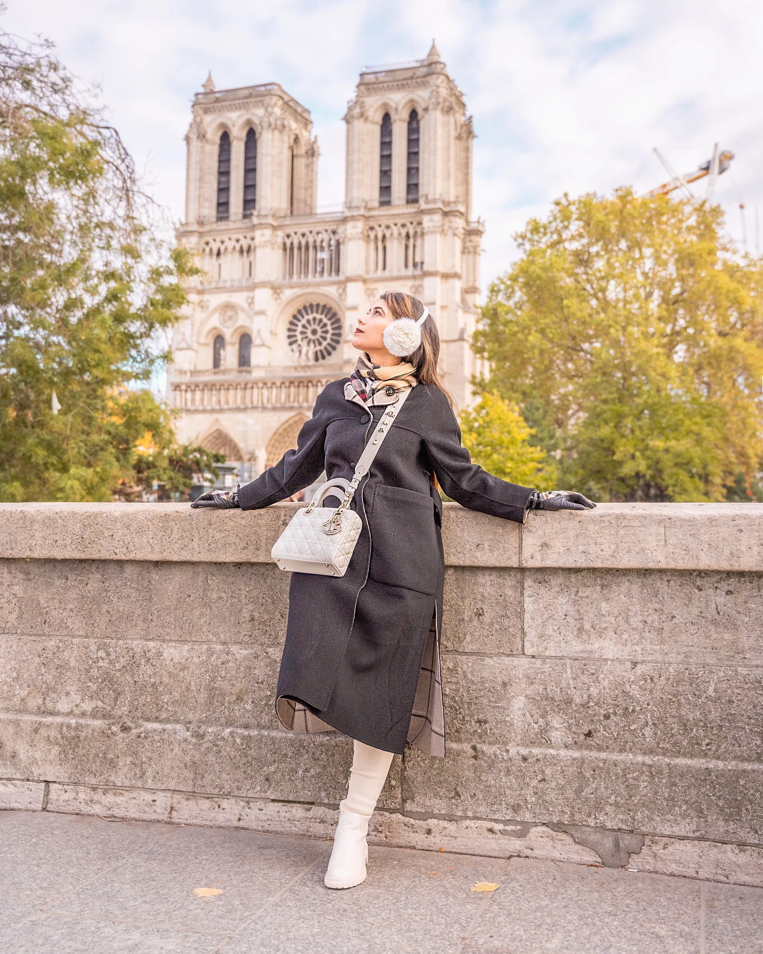 Woman in a black coat and earmuffs leaning on a stone balustrade with Notre-Dame cathedral in the background.