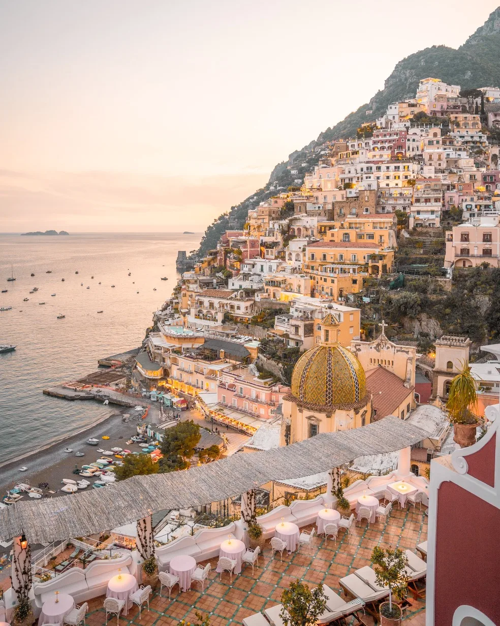 Sunset view over pastel cliffside houses and the tiled dome church of Positano overlooking the calm Amalfi Coast and a beach dotted with boats.