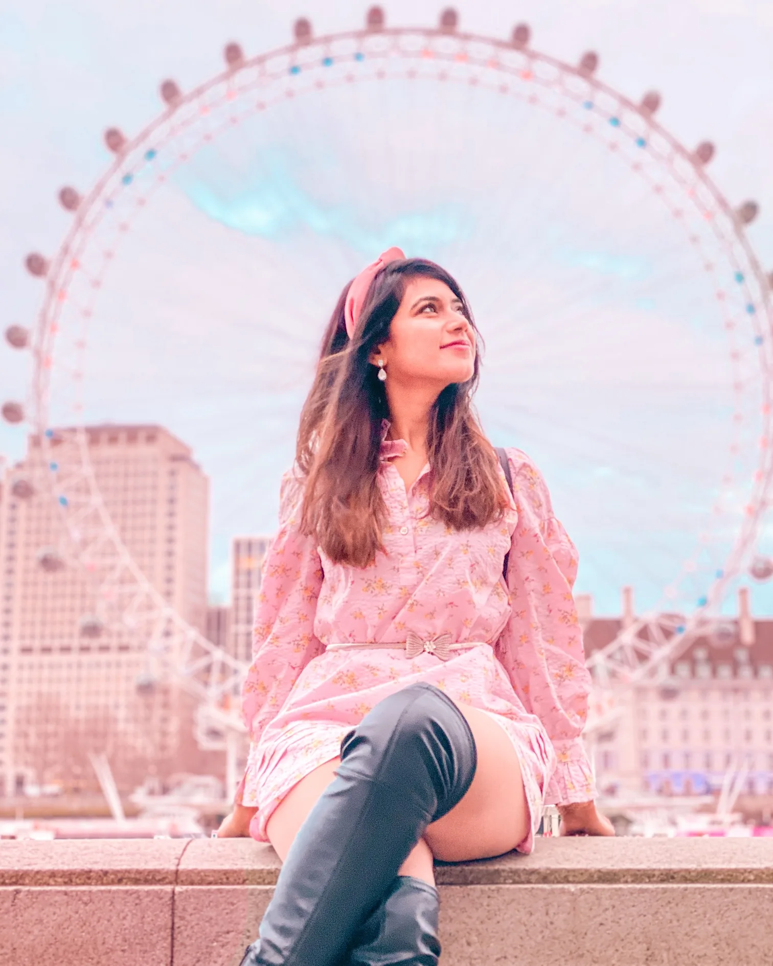 Young woman in a pink dress and headband sitting on a riverside wall with the London Eye ferris wheel blurred behind her