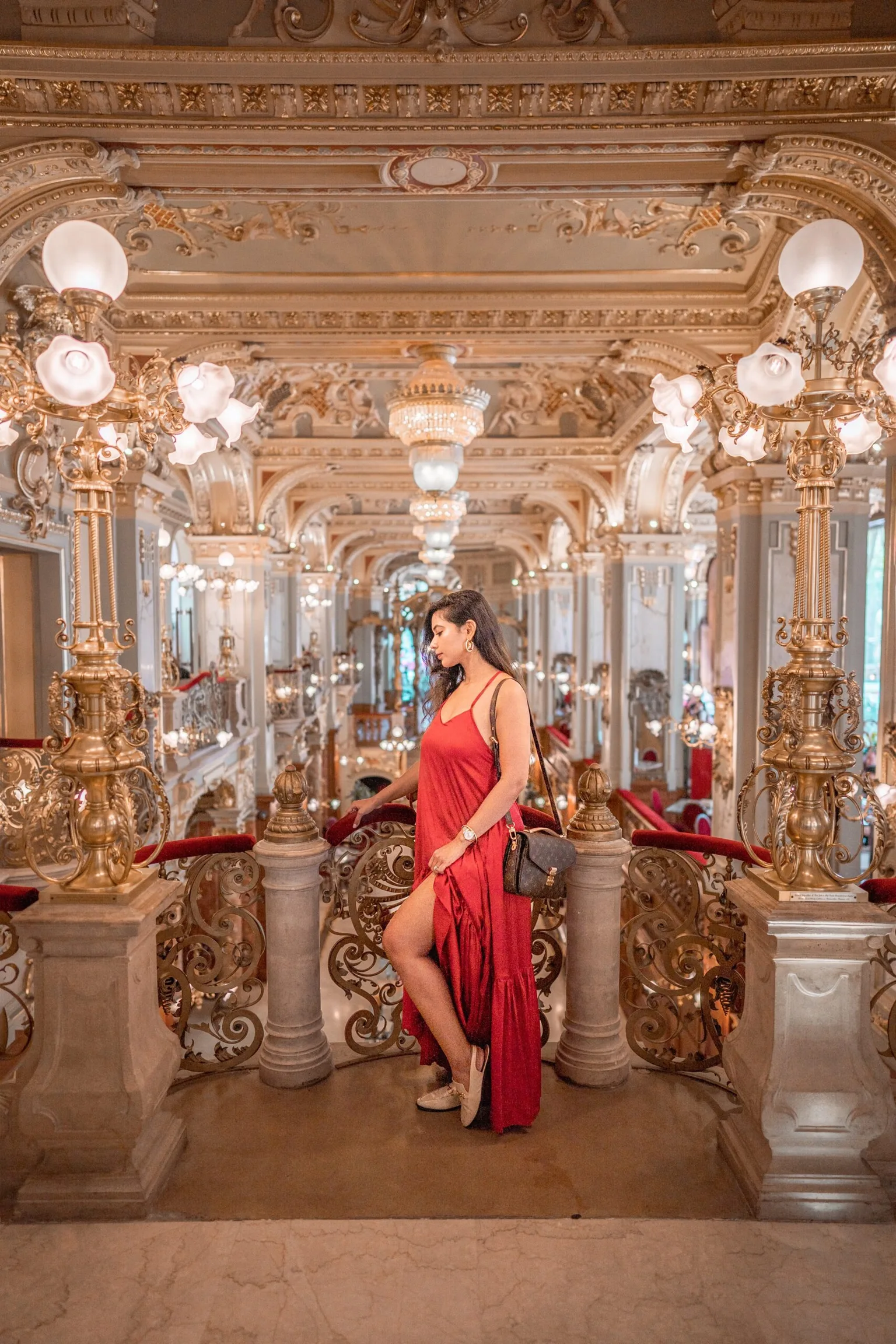 Woman in a flowing red dress posing on an ornate gilt balcony inside a grand, chandelier-filled palace interior
