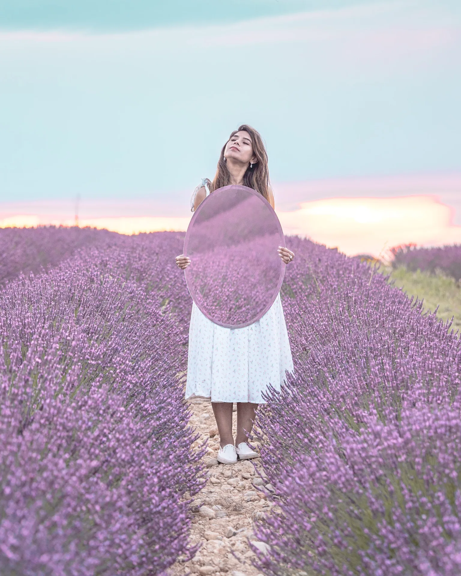 Woman in a white dress standing in a lavender field at sunset holding a round mirror reflecting the purple blooms.