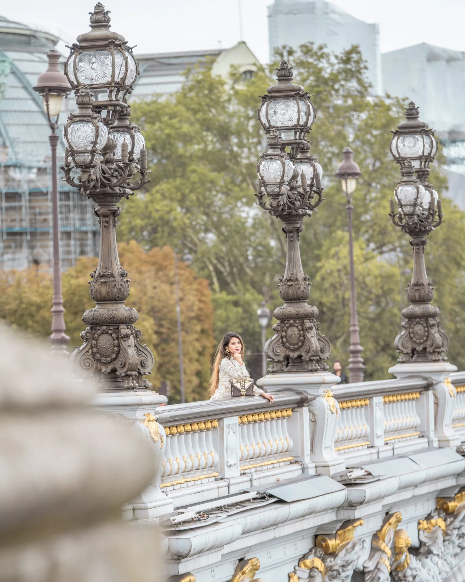 Woman in a patterned coat standing on an ornate Parisian bridge with decorative lampposts and gold accents.
