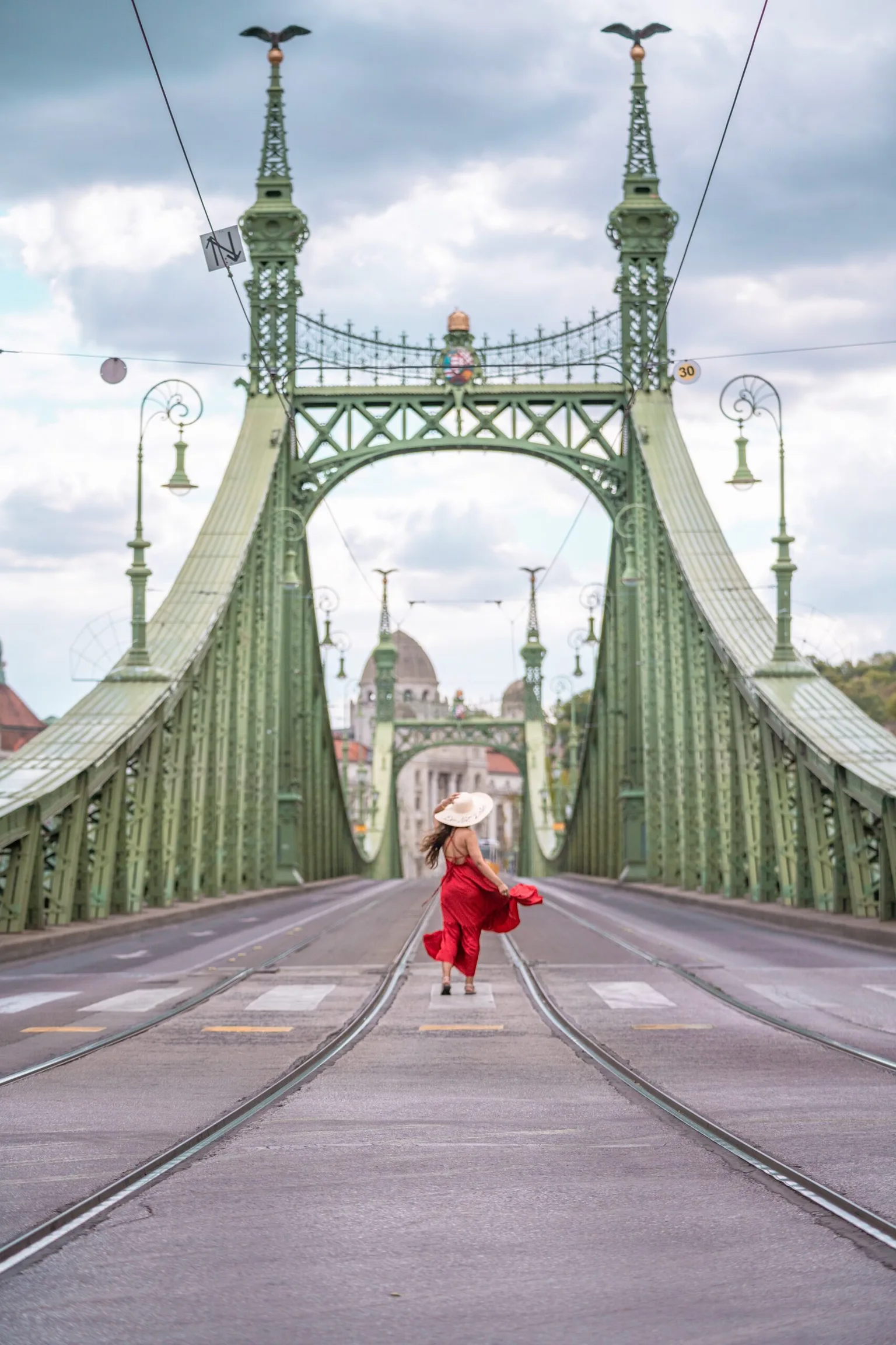 Woman in a flowing red dress and wide hat walking along tram tracks toward the ornate green Liberty Bridge.