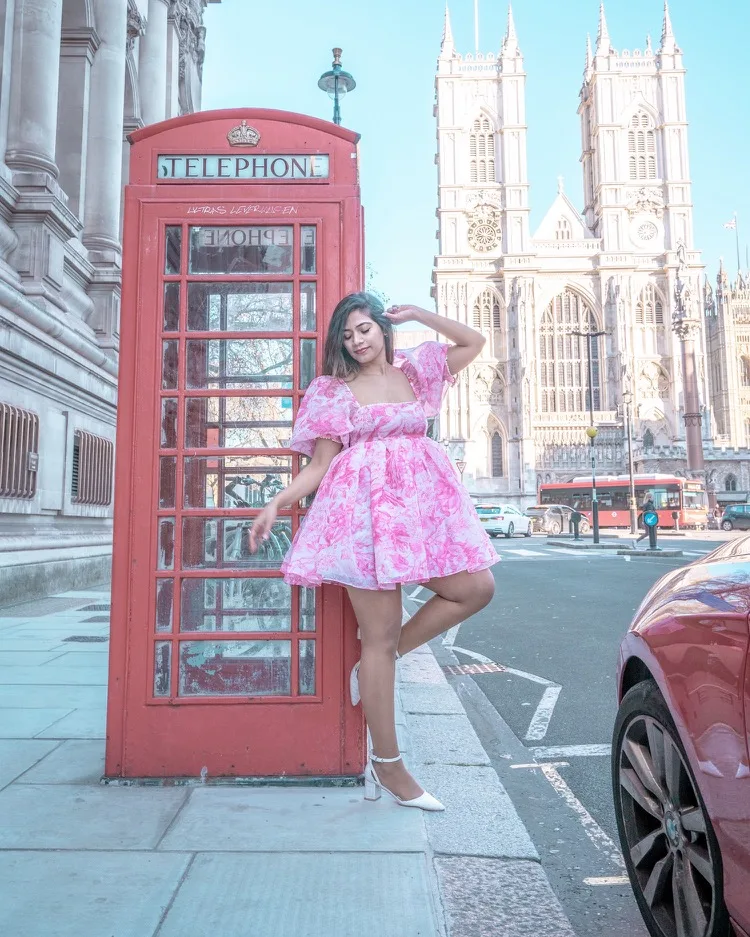 Woman in a pink floral dress posing beside a red London telephone box with Westminster Abbey in the background.