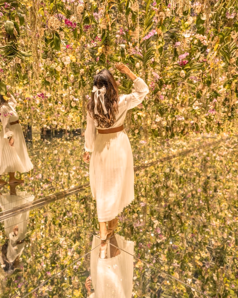 Woman in a cream dress stands in a mirrored flower installation surrounded by hanging blossoms