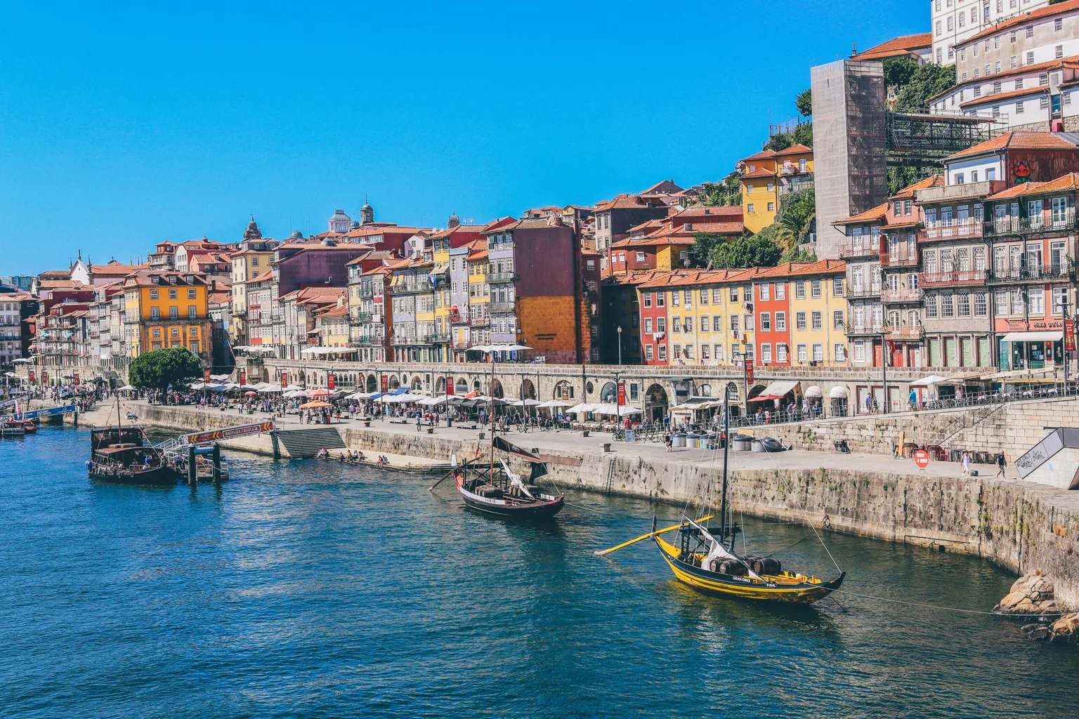 Colourful riverside buildings and cafés line the waterfront of Porto with traditional wooden boats moored on the blue Douro River.