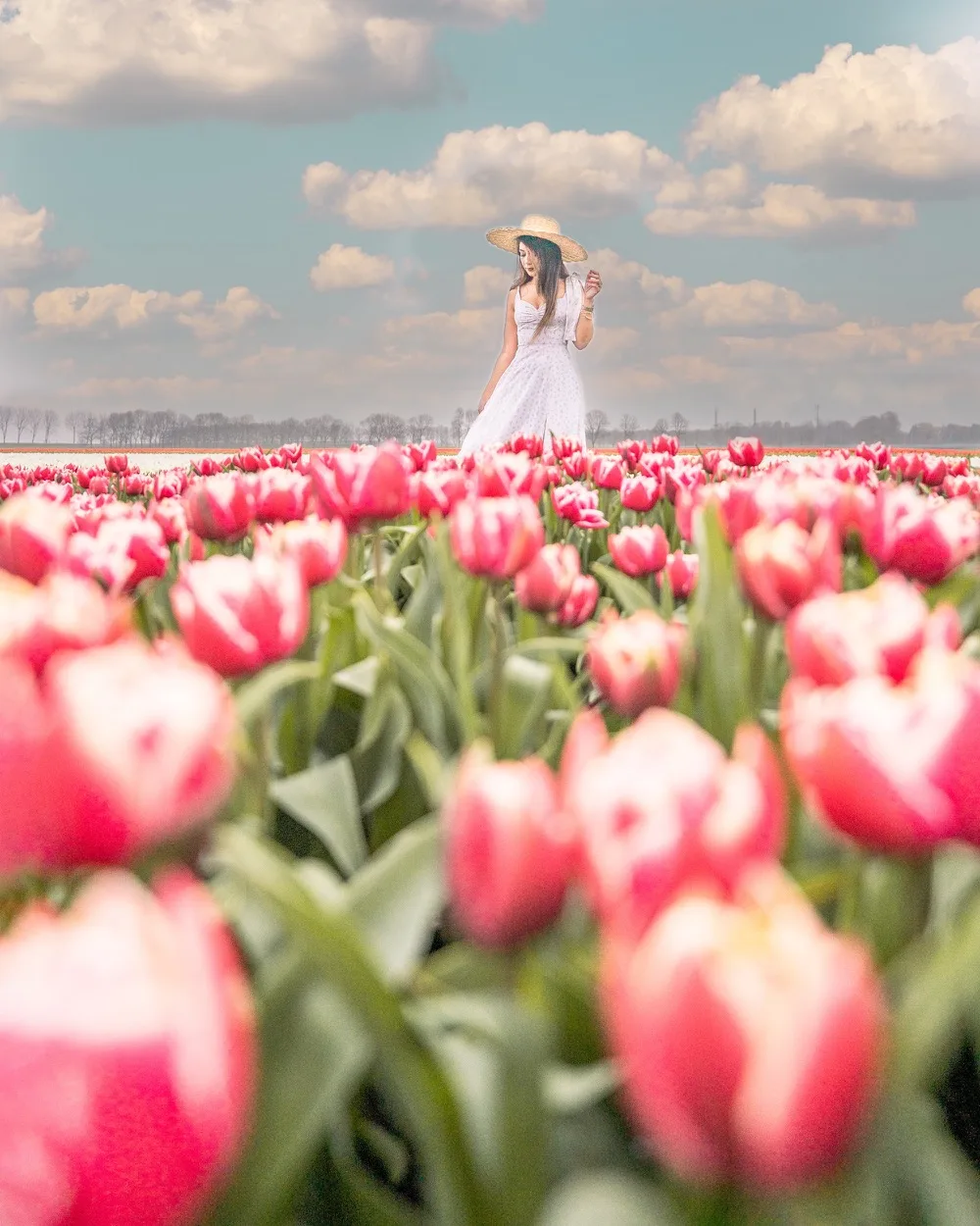 Woman in a white sundress and straw hat walking through a vast field of pink tulips under a blue sky with fluffy clouds.