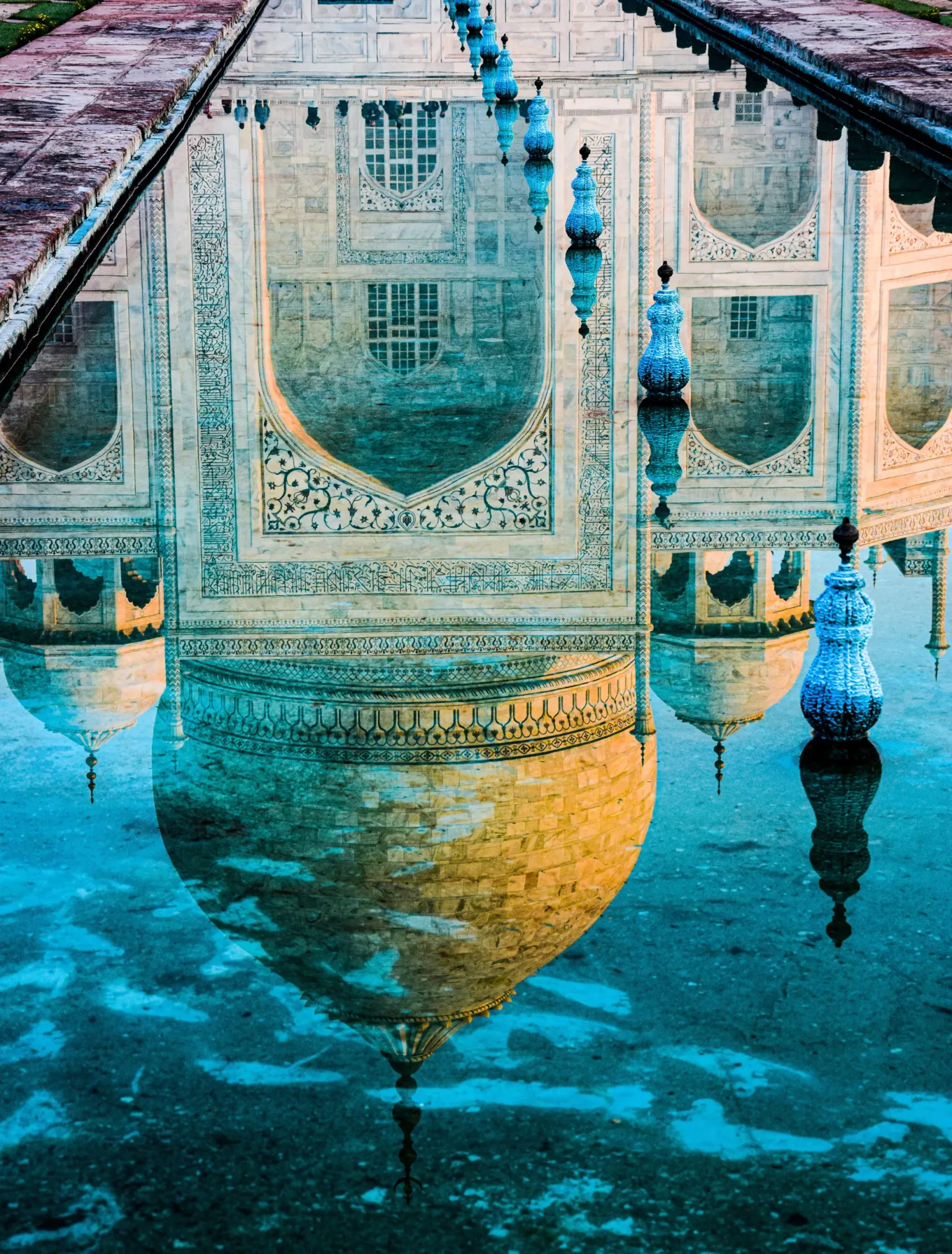Reflection of the Taj Mahal's ornate marble dome and archways mirrored in a blue-tinted pool with decorative finials.