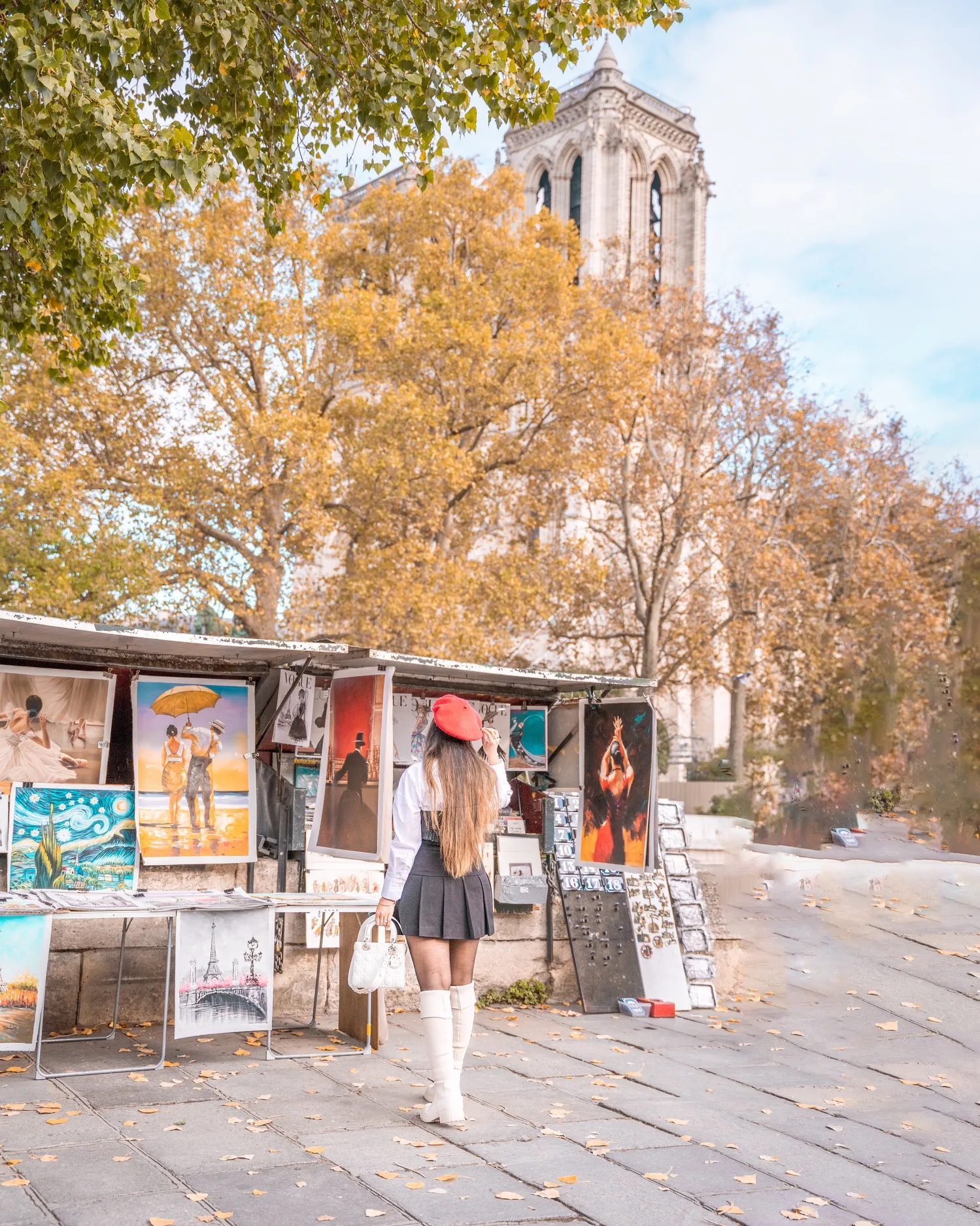 Woman in a red beret and white boots browsing outdoor art stalls with autumn trees and a cathedral tower in the background