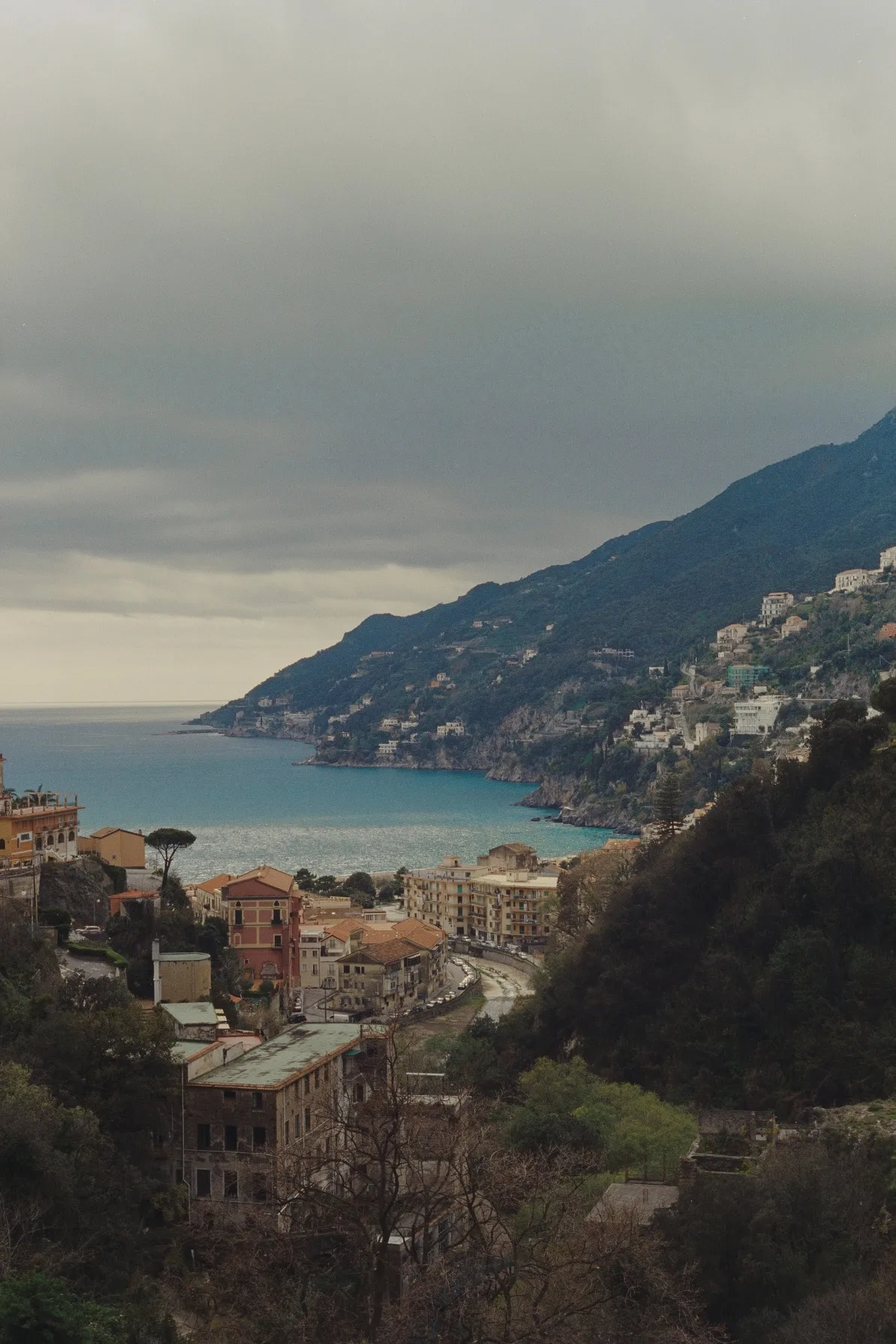 Coastal Italian village nestled between steep green hills and a calm turquoise bay under a cloudy sky.