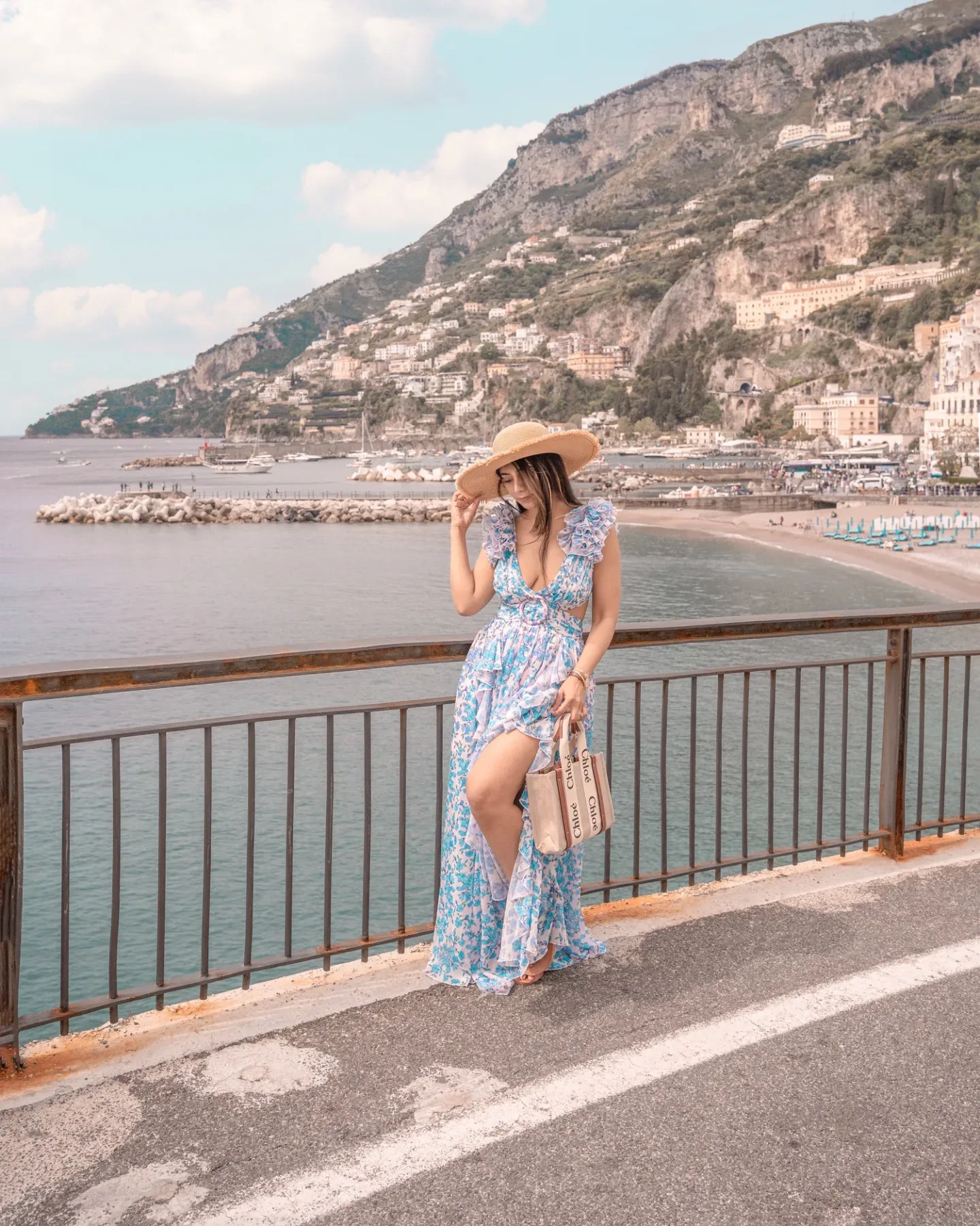 Woman in a blue floral sundress and straw hat leaning on a seaside railing with cliffs, villas and a marina in the background.